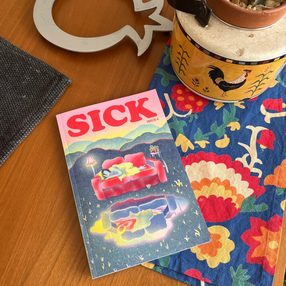 1: My copy of Sick magazine on the table, on top of a colorful floral dishcloth, next to a tea kettle with roosters painted on it and a trivet shaped like a pomegranate. The cover features a figure lying on a couch above the reflection of a figure lying on a couch, colors inverted.