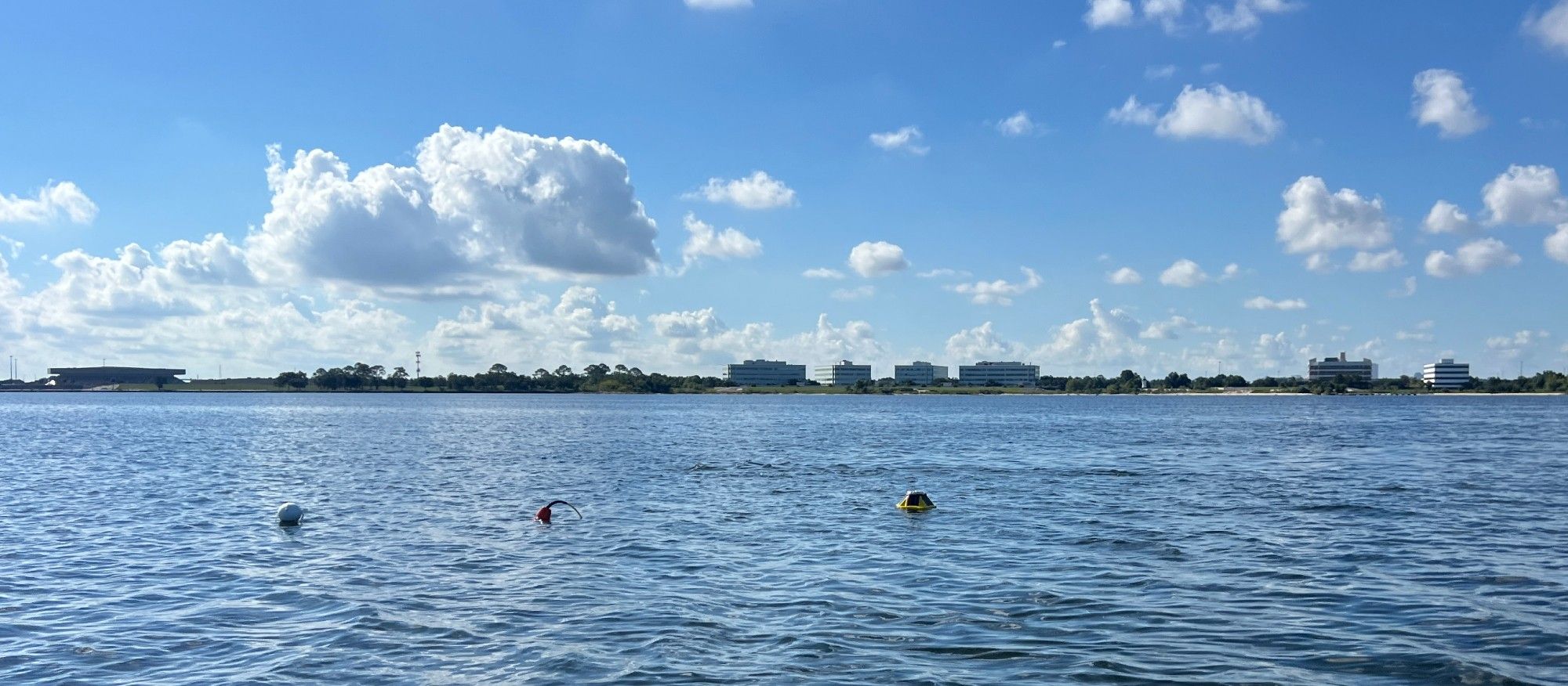 Image of a small yellow data buoy off the south shore of lake Pontchartrain, with the University of New Orleans' Research and Technology Park, Engineering Building, and Lakefront Arena visible in the background