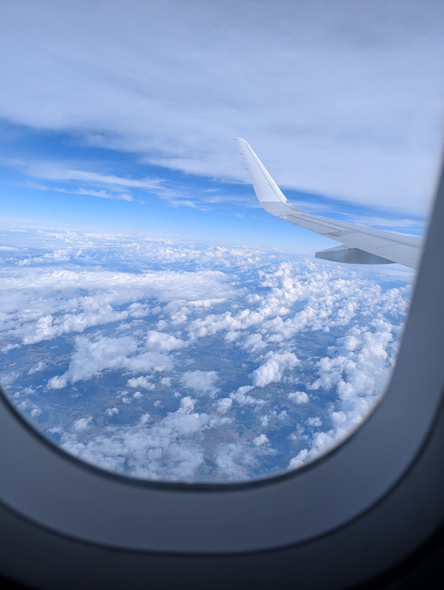 Blick aus einem Flugzeugfenster. Man sieht eine Wolkendecke und ein Stück Flügel.