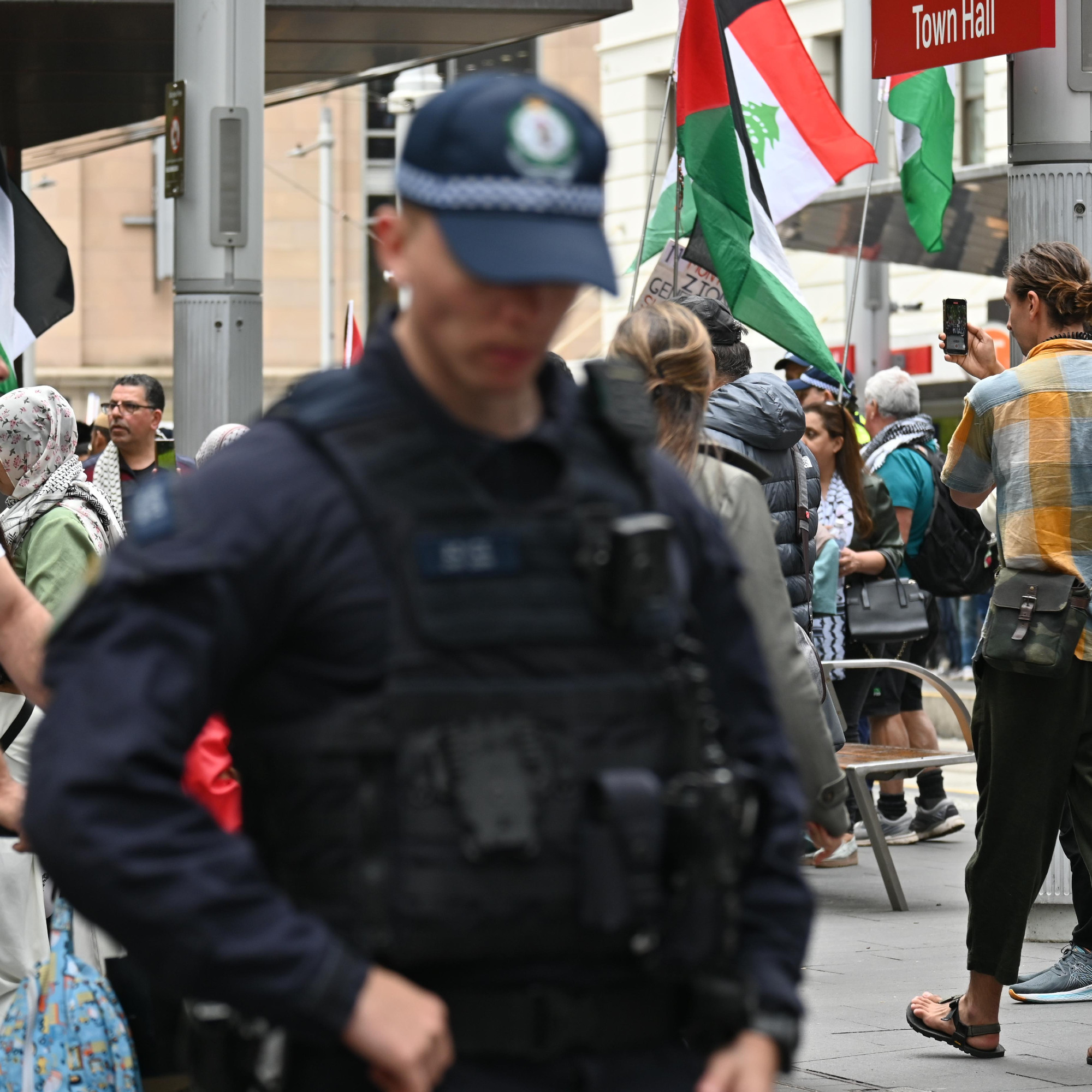 A police officer, out of focus, with his head tilted down, and hands resting on his toolbelt. Behind him are Palestinian and Lebanese flags flying at a protest