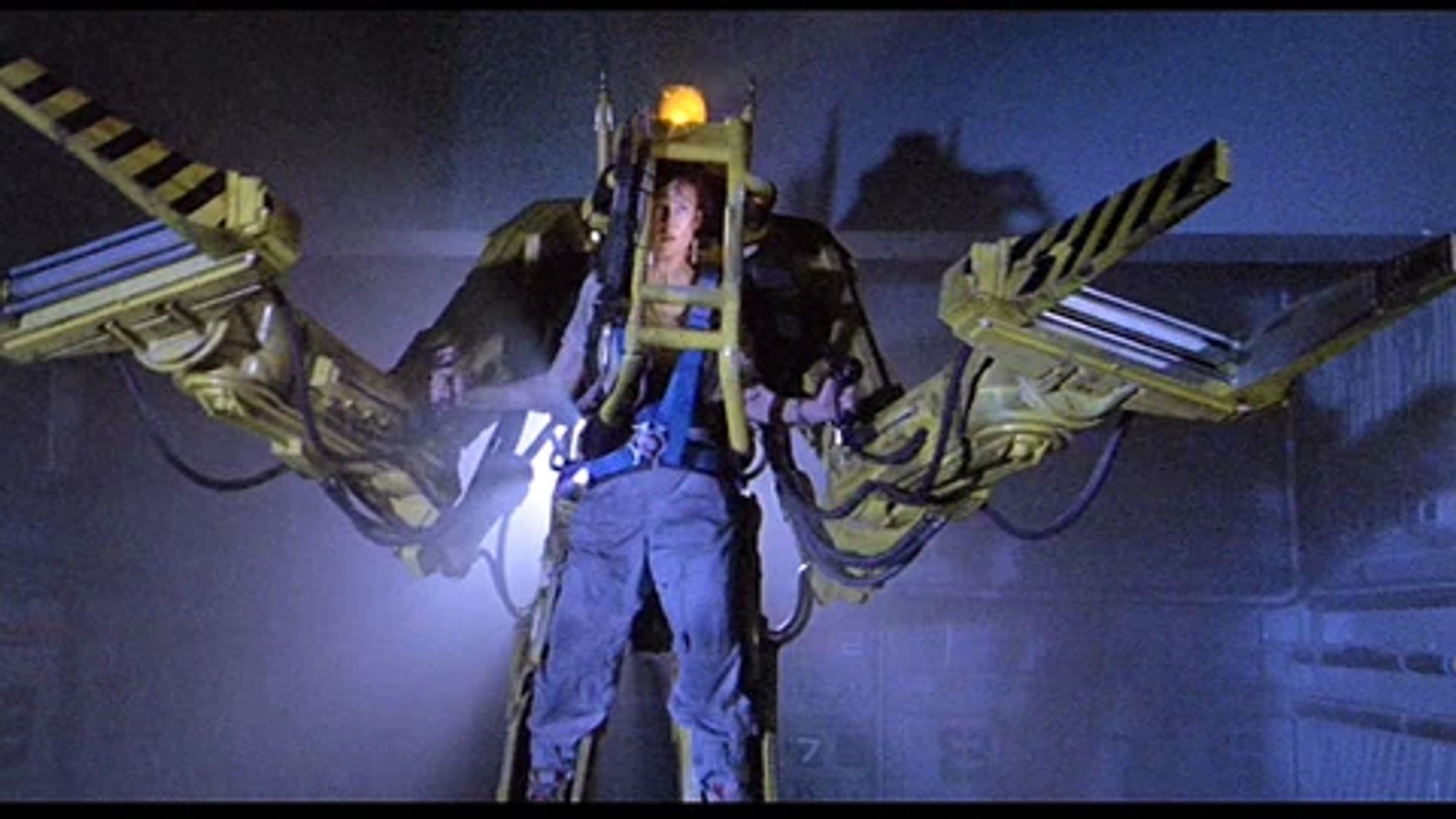 Still from the 1986 movie Aliens, with Ellen Ripley preparing for battle against the Xenomorph Queen. She wears a simple shirt and jeans, and stands and wears the external P–5000 Powered Work Loader mechanised exoskeleton. Its clamp prongs on her arms are menacing out to the viewer.