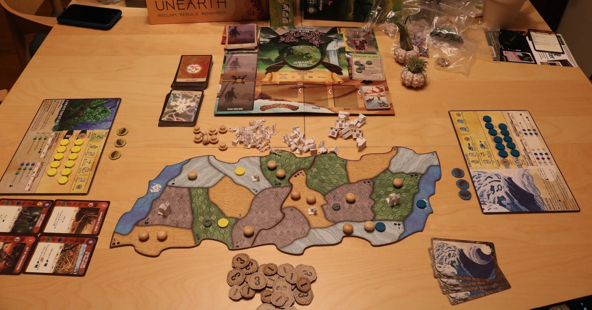 A table setup for two players to play Spirit Island on a wooden table. Two connected boards with colour and patterns demarking wetlands, mountains, deserts, and jungles, are covered with little huts, figures of towns and cities and conquistador-like figurines. A side board and some player cards with playable power cards surround the playing field.