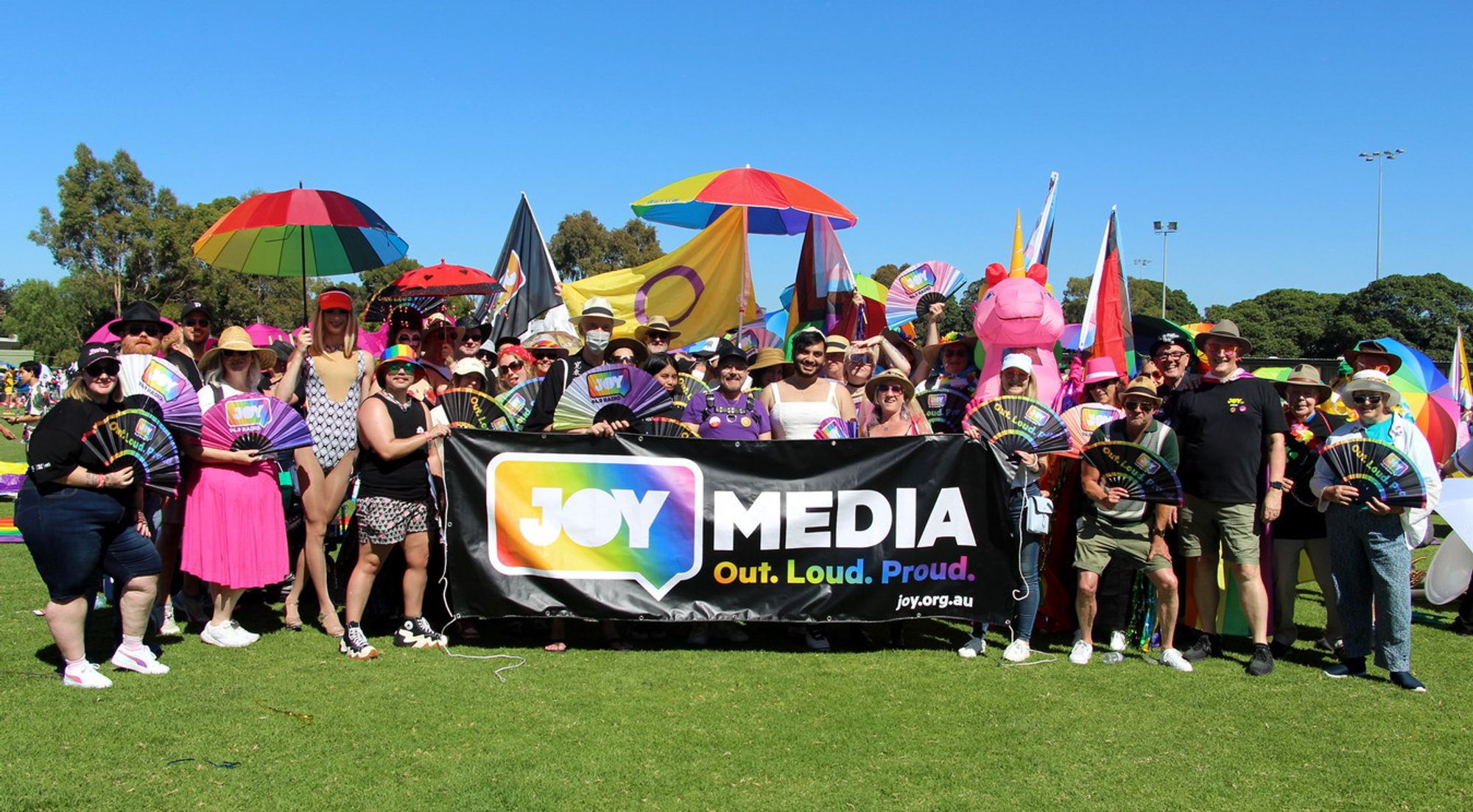 A group photo of the fifty or so people marching with JOY at Melbourne's Midsumma Pride March this year. The colours are super vivid. The atmosphere is joyful. The grass is green, and fittingly, the sky is blue.