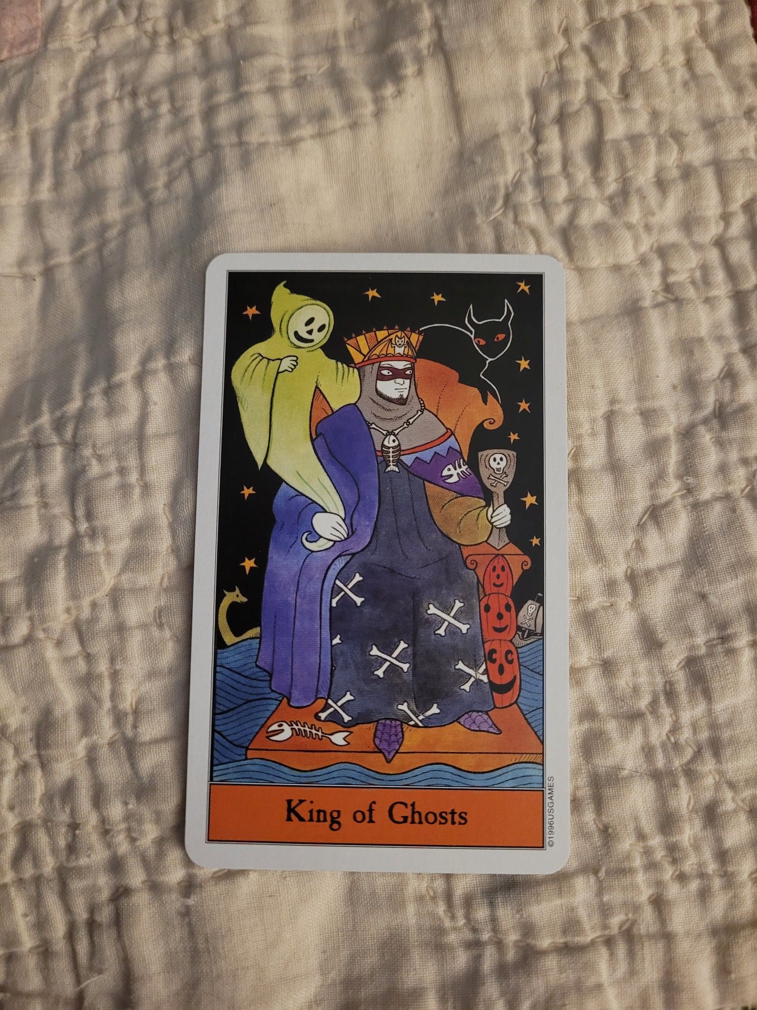 King of Ghosts card in the Halloween Tarot, by Karin Lee and Kipling West. Equivalent to the King of Cups in traditional decks, this king's robe is decorated with crossed bones and fish skeletons. He has a friendly ghost looking over one shoulder and a black cat on the other.