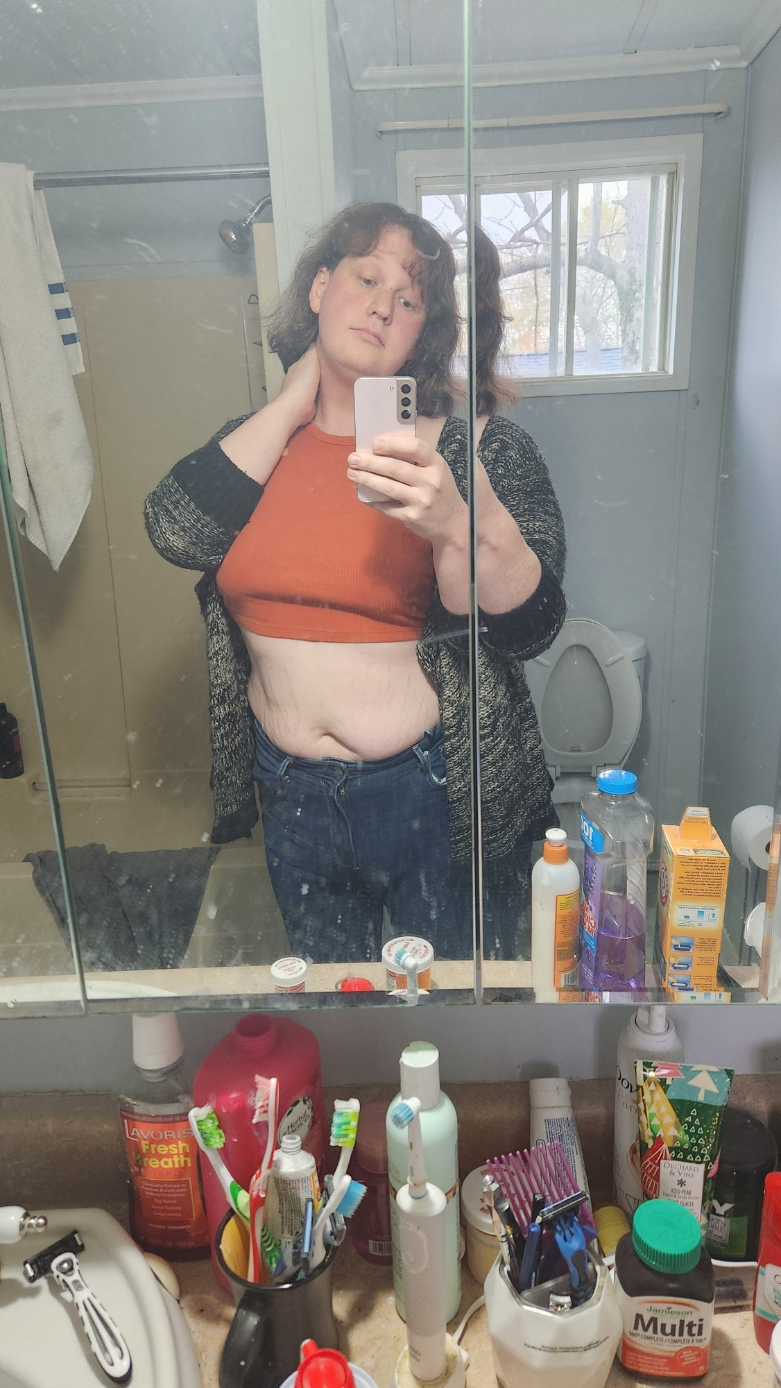 A brown haired woman taking a mirror selfie, she is wearing blue jeans, a burnt orange crop top, and a beige and black cardigan.