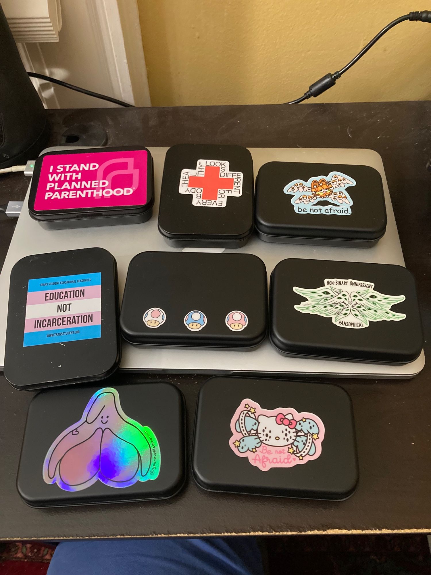 8 black tins with stickers on them, including a holographic clitoris cartoon and various biblically accurate angel depictions.