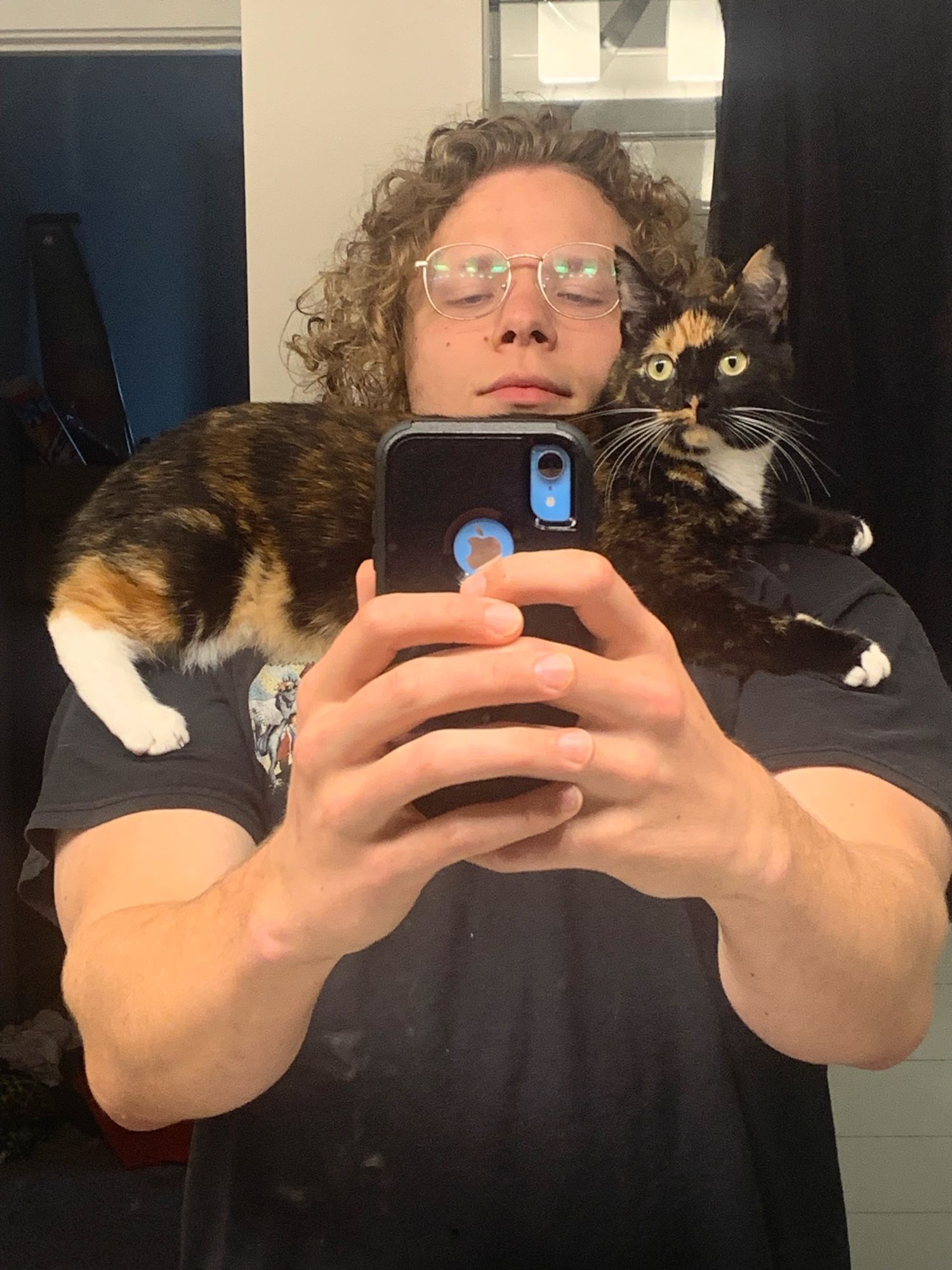 Young man with long curly hair with a cat draped over his chest and shoulders, while he takes a selfie.