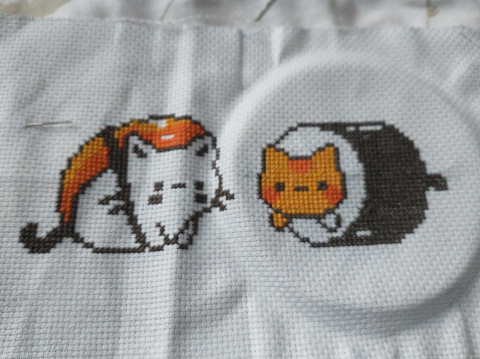 A piece of completed cross-stitch of two cats designed to look like sushi. The cat on the left is white and looks like a piece of salmon sashimi. The cat on the right is ginger and looks like a maki roll.