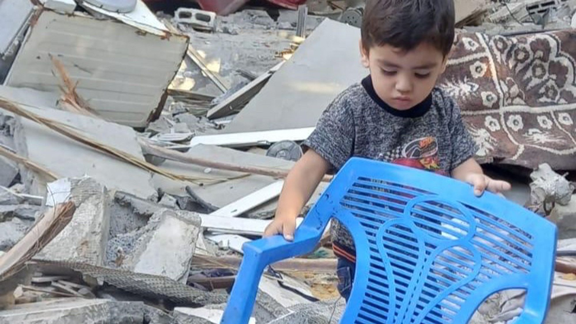 in the middle of rubble left from broken house, small child moving a blue plastic chair around, in the background broken pieces of beton and fabric laying around