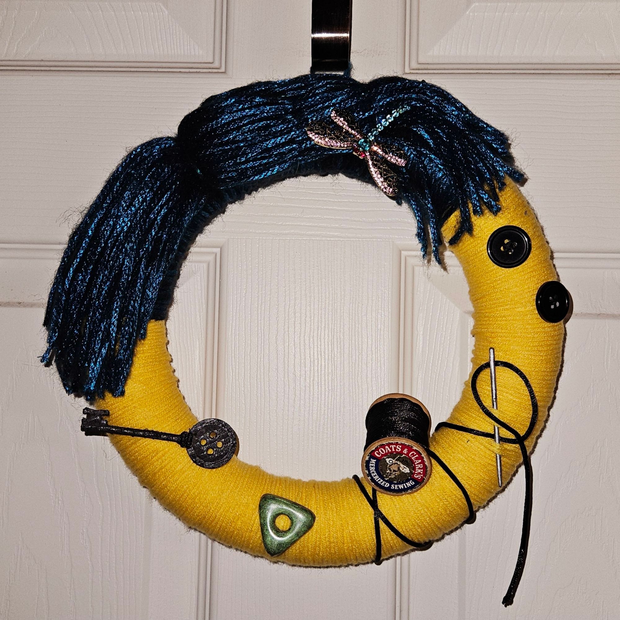 Image is of a small diameter homemade wreath themed from the character Coraline. 3/4 of it is yarn wrapped in bright yellow like her raincoat, and the rest is yarn wrapped in a shiny teal like her hair. There is a rhinestone dragonfly hair clip in the hair, and on the yellow part are two black buttons, it wouldn't spool with black thread going through a large silver needle, a green triangular stone with a circle hole in the middle, and a skeleton key with a button on top.