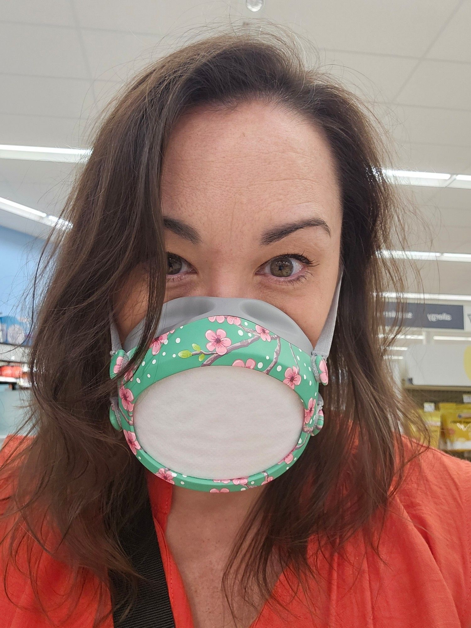 Image is a middle aged woman from the shoulders up. She has shoulder length dark brown hair, hazel eyes, and is wearing an orange shirt. Half her face is covered with a silicone seal Flo Mask with a pale green and pink cherry blossom print on the frame.