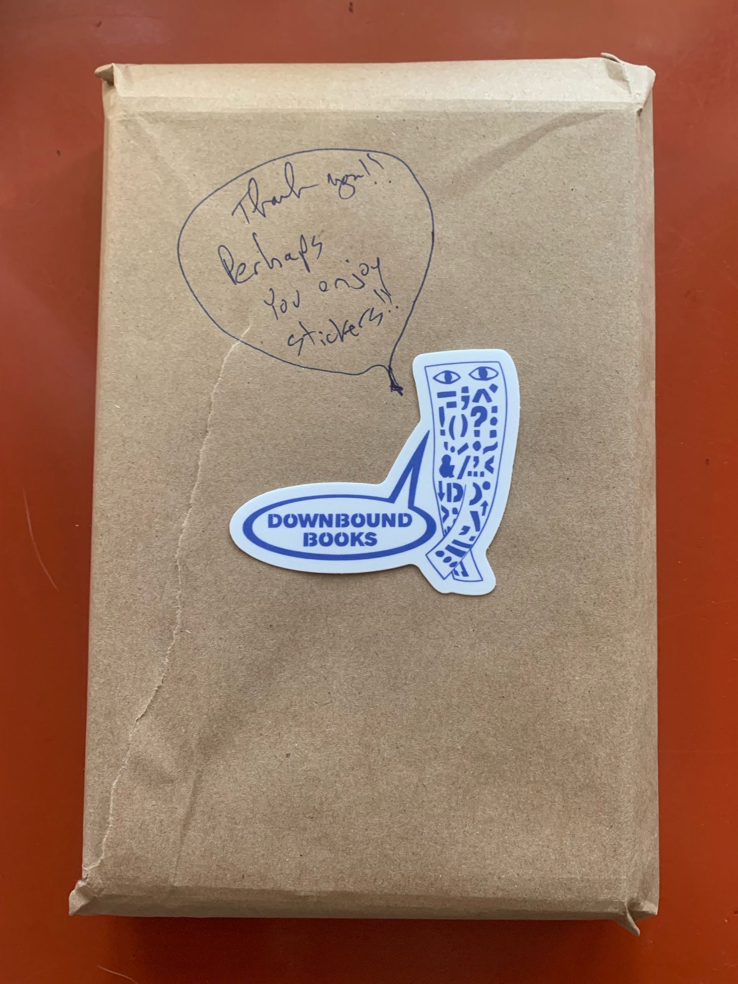 A parcel wrapped in brown butcher paper. On it is a sticker of an oblong cartoon character standing with its legs crossed at the ankles and a speech bubble that says “Downbound Books.” On the paper is drawn, in pen, another speech bubble, this one saying, “Thank you! Perhaps you enjoy stickers!!”