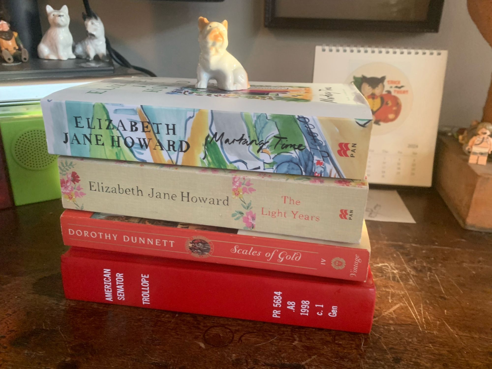 A stack of four books topped by a small ceramic dog: Trollope’s The American Senator, Dorothy Dunnett’s Scales of Gold, and Elizabeth Jane Howard’s first two Cazalet Chronicles books, The Light Years and Marking Time.