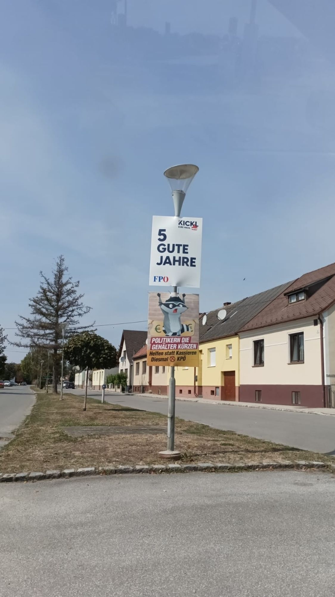 An einer Lampe hängt oben das Plakat der FPÖ mit "5 gute Jahre!" und darunter unser Plakat mit dem süßen Waschbären mit "helfen statt kassieren"