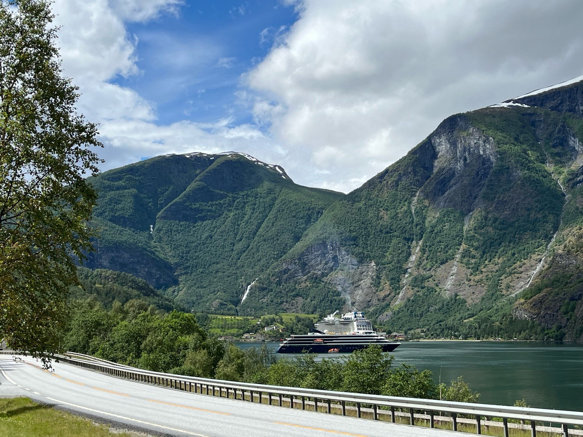 Blick auf einen Fjord, im Vordergrund führt eine Straße am Ufer entlang, im Hintergrund Berge, darüber ein blauer Himmel mit weißen Wolken. Im Wasser liegt ein Kreuzfahrtschiff.