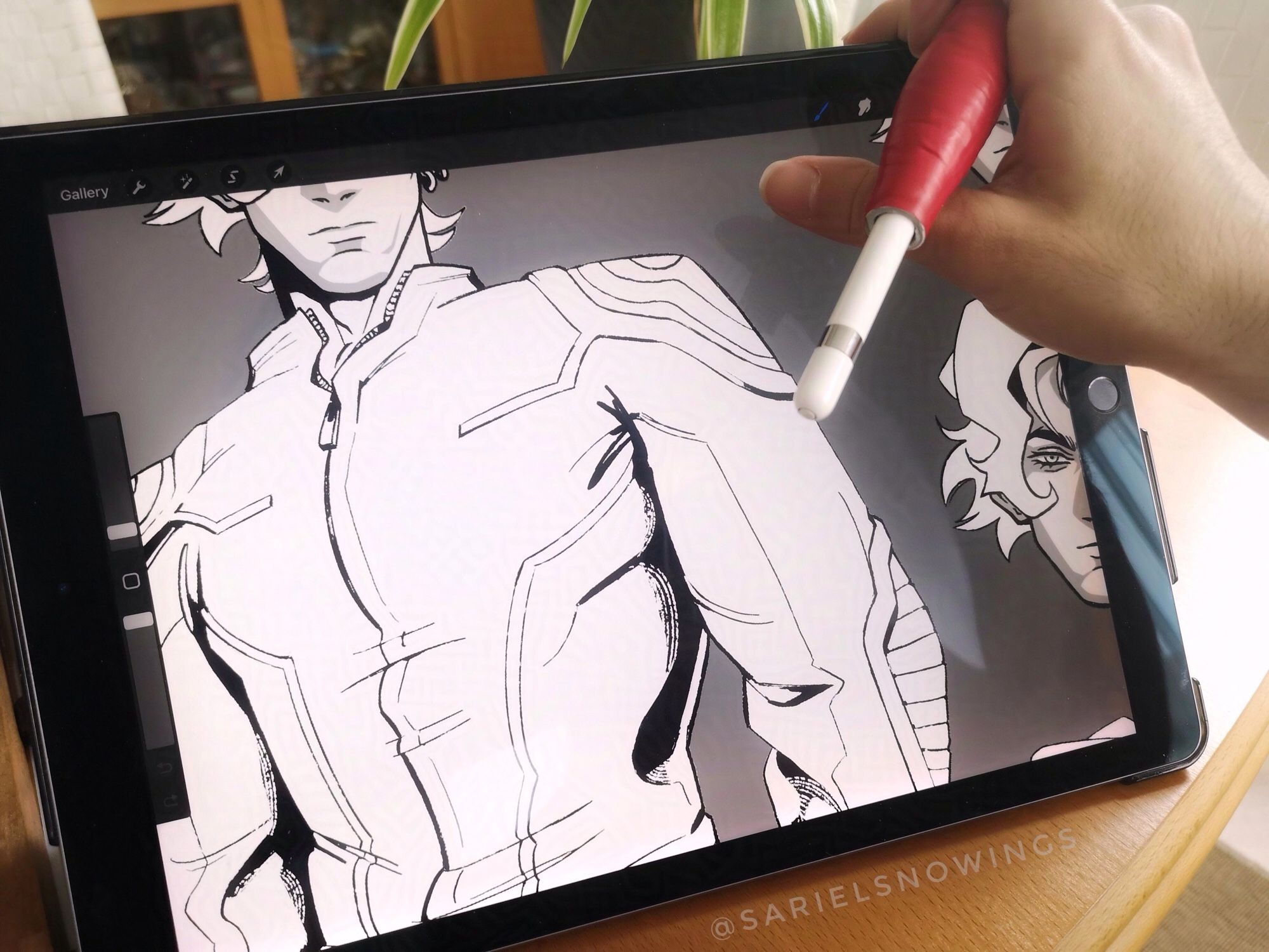 A picture of Sariel’s tablet on a wooden table. The screen displays a zoomed-in view of a drawing in progress. It features a male character wearing motorcycling gear inked in Sariel’s usual style. We can only see his torso and the bottom half of his face. On the right side of the screen there’s also a cropped drawing of the character’s face with a different expression. Sariel’s hand is also in the picture, resting on the top right corner of the tablet and holding their Apple Pencil.