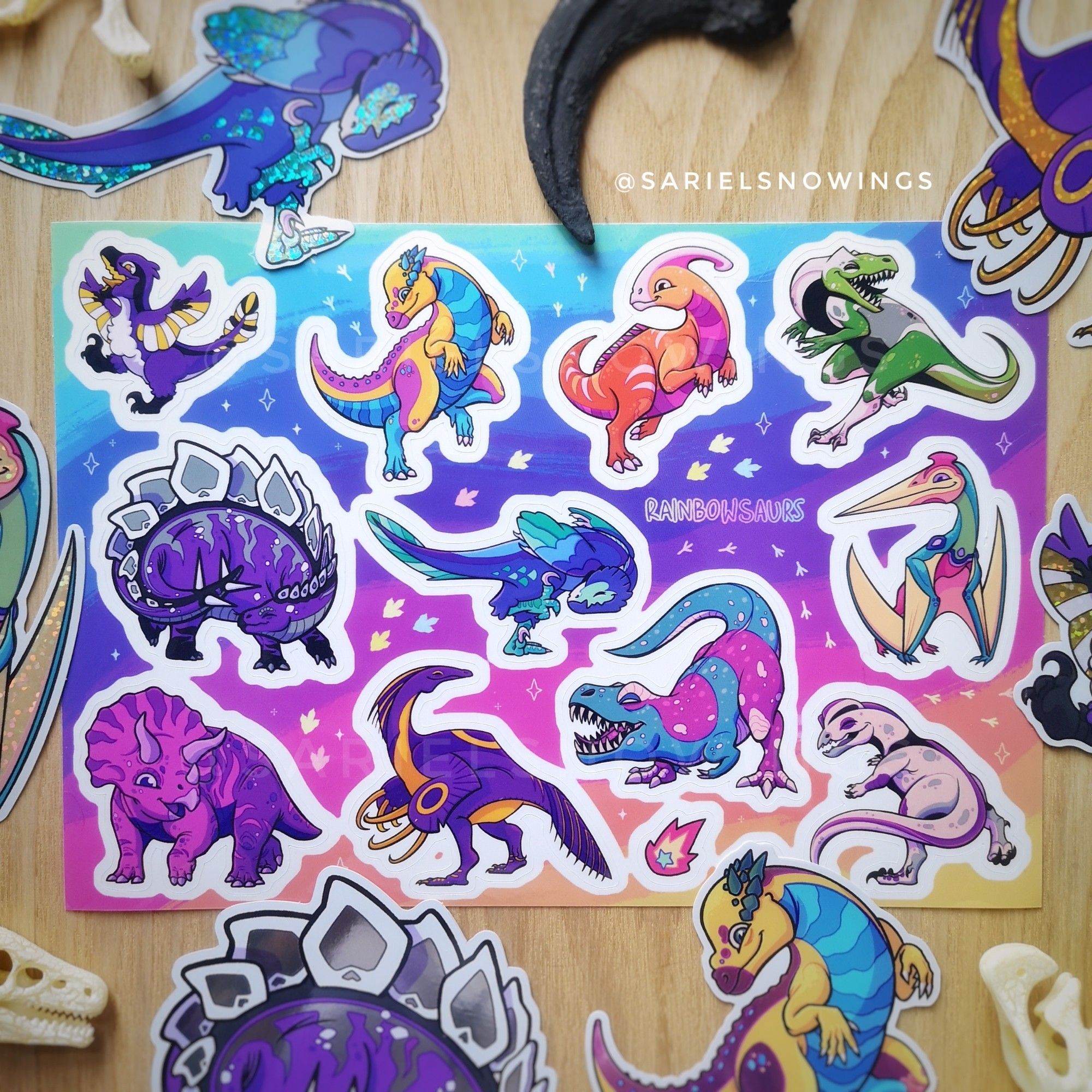 A picture of a sticker sheet on a wooden table. The sheet features a variety of “Rainbow-saurs”, dinosaur drawings, each with the colour palette of a pride flag. They’re also puns mixing the queer identities with the name of each dinosaur. Top to bottom, left to right, they are the following:
ENBYlociraptor: non binary (enby)   velociraptor
PANchycephalosaurus: pansexual   pachycephalosaurus
ParaSAPPHOlopus: lesbian (sapphic)   parasaurolophus
AROsaurus: aromantic   allosaurus
ASEXtegosaurus: asexual   stegosaurus
GAYnonychus: gay   deinonychus
TRANSnnosaurus Rex: transgender   tyrannosaurus rex
QUEERtzalcoatlus: queer   quetzalcoatlus
BIceratops: bisexual   tricetratops
INTERizinosaurus: intersex   therizinosaurus
DEMIlophosaurus: demisexual   dilophosaurusAround the main sheet there are bigger sticker versions of some of th