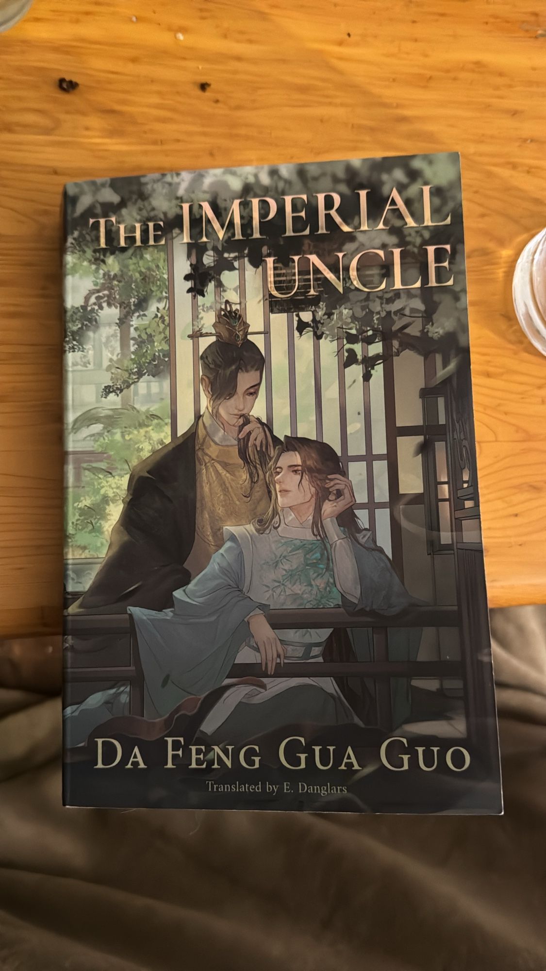 The Imperial Uncle Book Cover by author Da Feng Gua Guo and translated by E. Danglars featuring two men sitting by a wooden fence one man wearing a blue robe is leaning on the fence while the other man sits behind him bringing a lock of hair towards his lips.