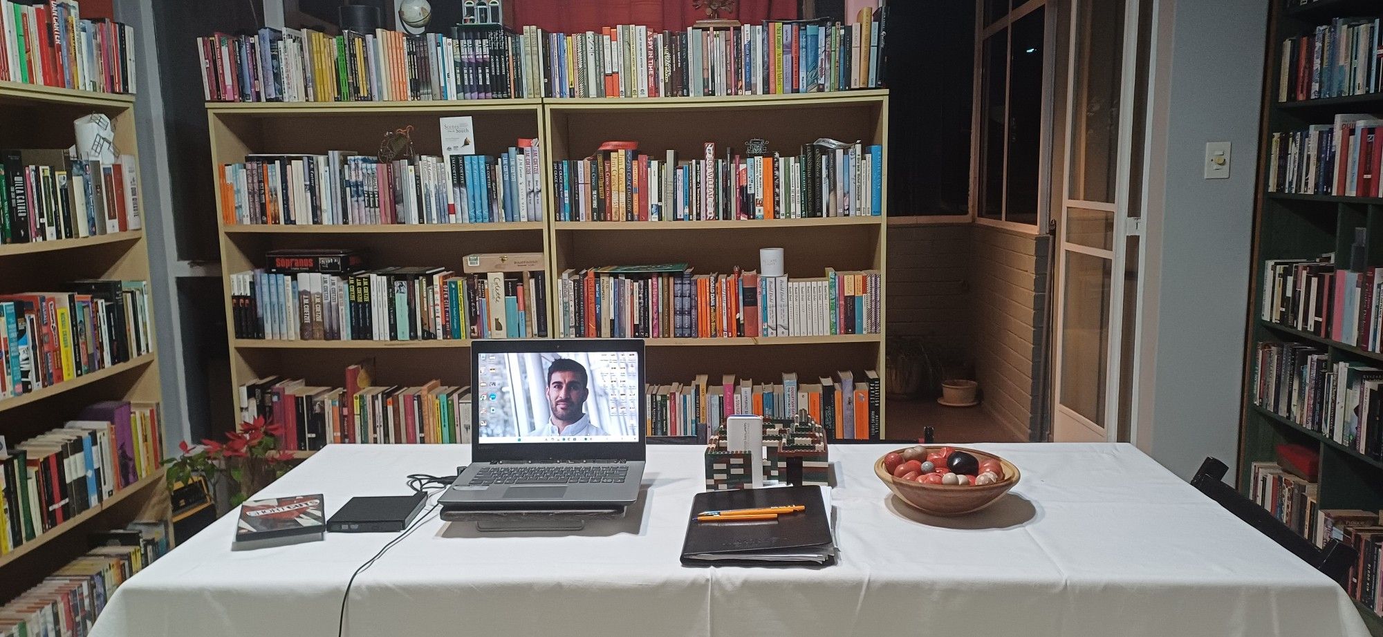 Lapyop and external DVD player resting on a table covered with a white cloth; also on the table a toy brick house, a bowl of soapstone hearts, a folder of paper and some ballpoint pens, as well as the DVD cover for Robert Altman's "Short Cuts". Behind the table and on the left and right of the frame are bookcases filled with fiction (alphabetised by author surname) and a door leading to the room where the fourth photograph was taken.