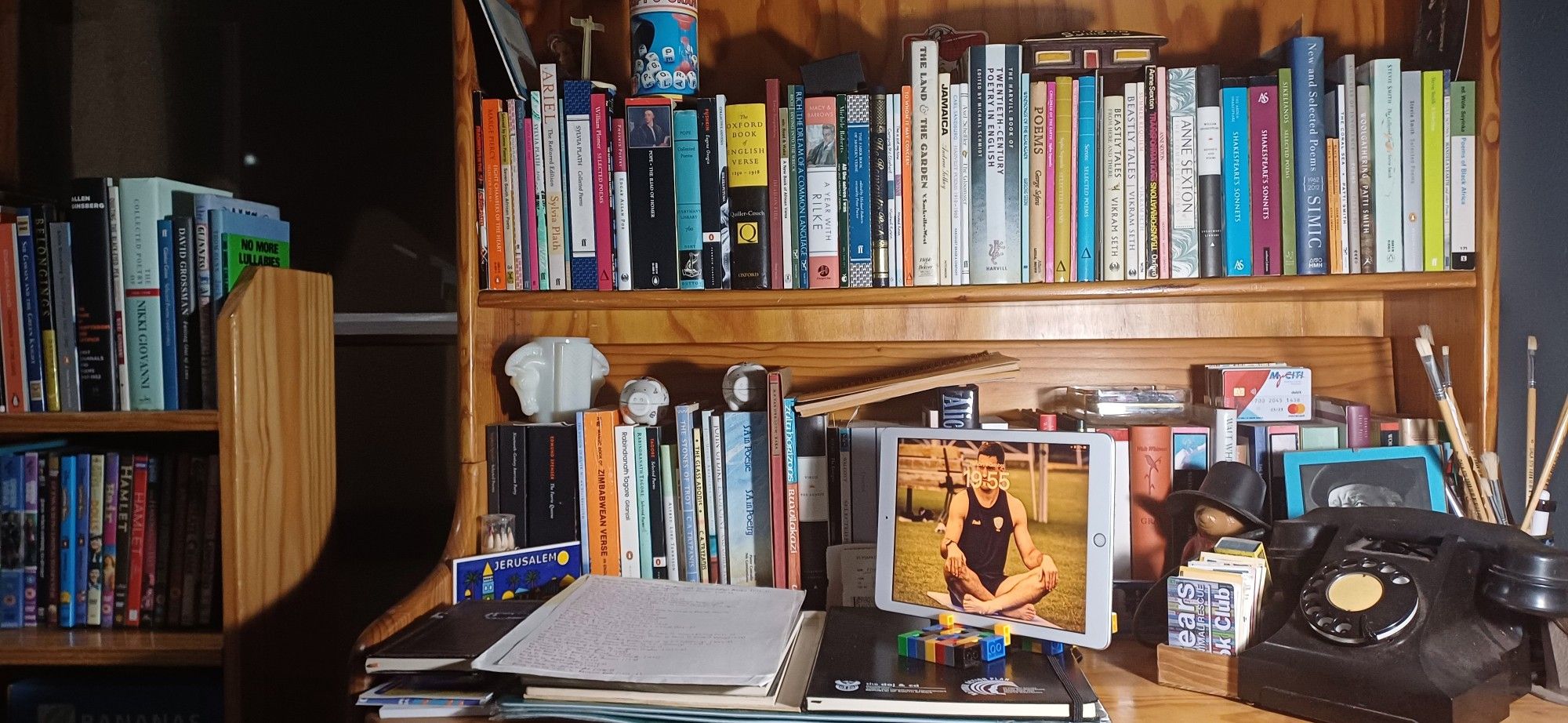 A cluttered upright wooden student's desk houskng shelves of poetry; on the desk itself can be seen an iPad, a.1950s Bakelite telepjone, paint brushes, a Paddington money box, and several notebooks.