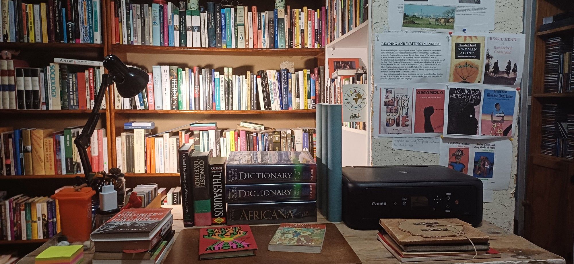 A 1952 raw wood table serving as a workint desk with an anglepoise lamp, dictionaries, subdry books, notebooks' and a printer; behind the desk are bookcases of non-fiction (alphabetised by author surname) and a noticeboard.