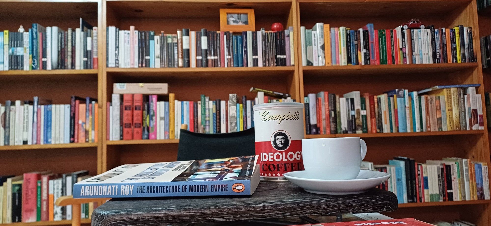 Arundhati Roy's "The Architecture of Modern Empire" next to a cup of espresso and a can of coffee beans styled like Campbell's soup can but with an image of Che Guevara and labelled 'Ideology Coffee' in front of 3 shelves of fiction alphabetised from H through K, an empty Cuban cigar box,  a framed photograph, and a model lf a red VW Beetle.