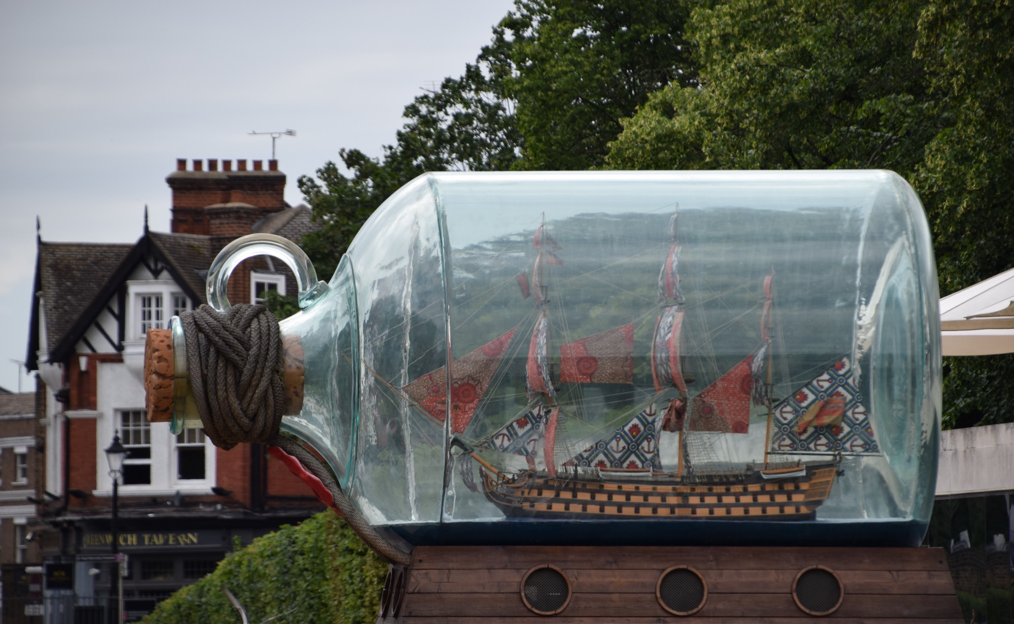 A oversized bottle with a sailing vessel inside is on display. The vessel has colorful sails, with red the dominant color. The bottle has a cord braided around its neck and a large cork as its stopper. Behind the bottle are trees to the right and an old Tudor-looking tavern to the left. The sky is gray and overcast.