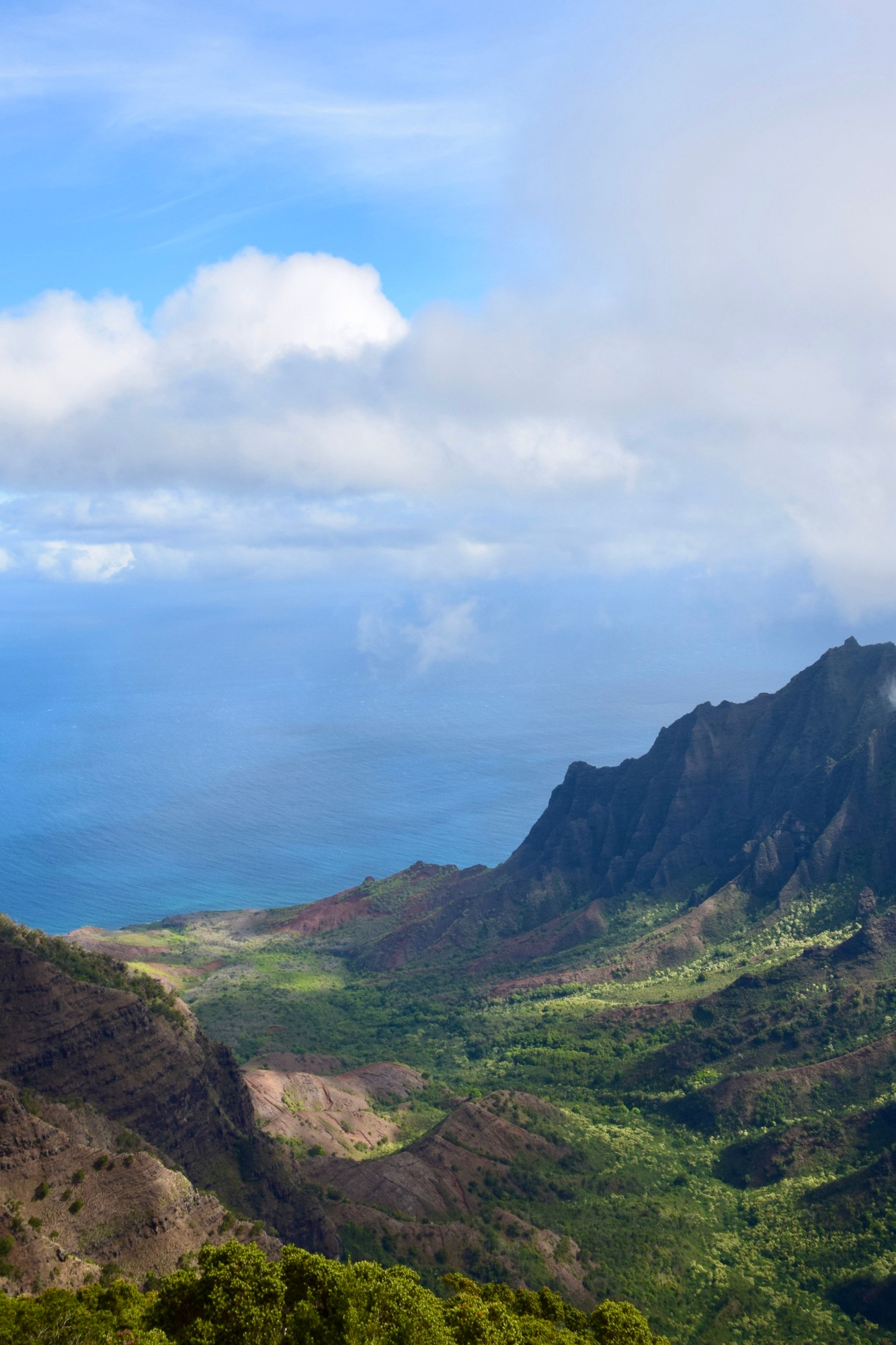 Craggy hills lead to a valley overlooking a misty blue ocean with a blue sky and white clouds above. 