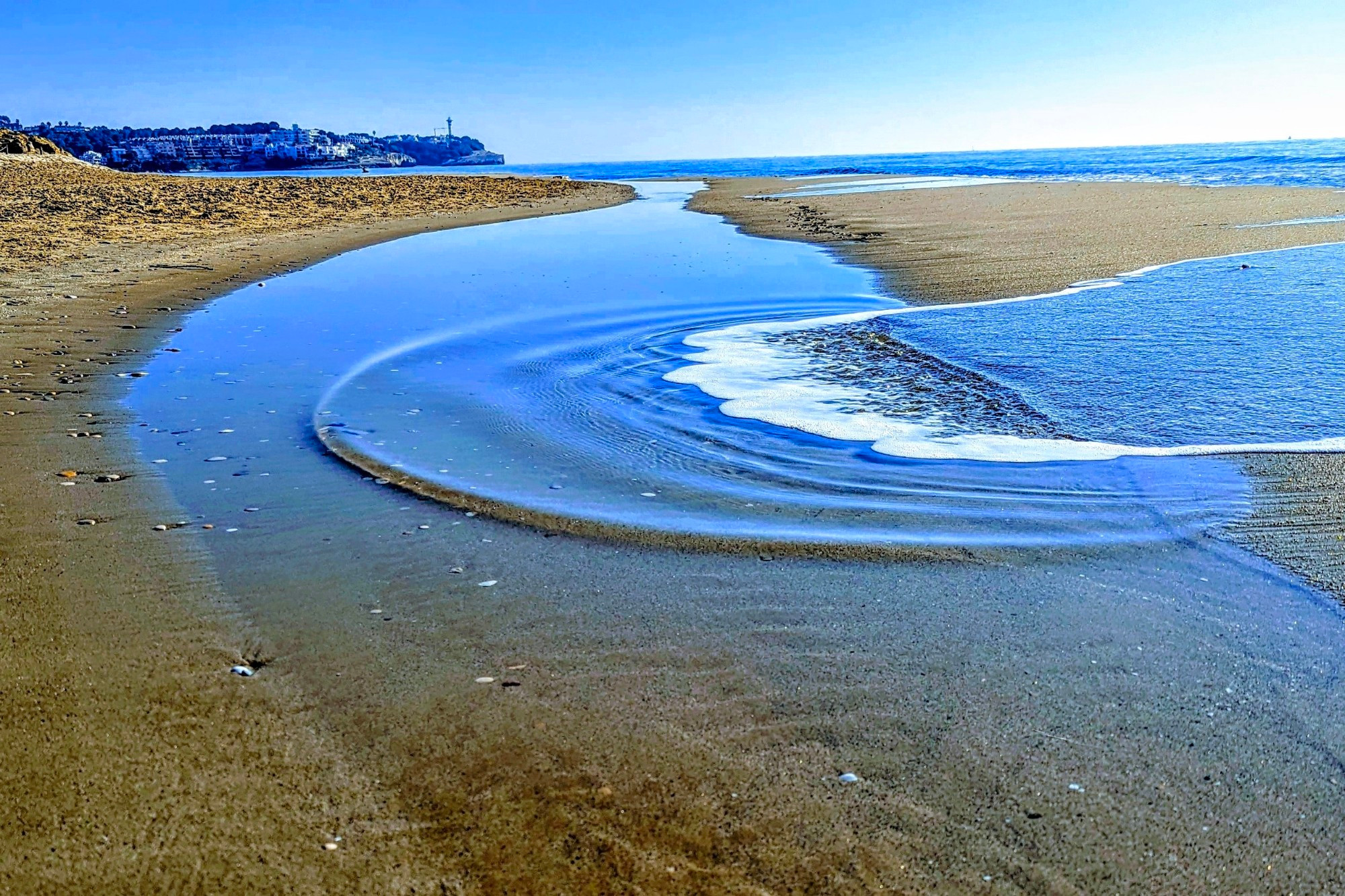 Ein schmaler Wasserlauf schlängelt sich durch den Sandstrand und mündet in das ruhige Meer. Im Vordergrund sind kleine Wellen und Schaumkronen zu sehen.


A narrow waterway winds its way through the sandy beach and empties into the calm sea. Small waves and foam crests can be seen in the foreground.