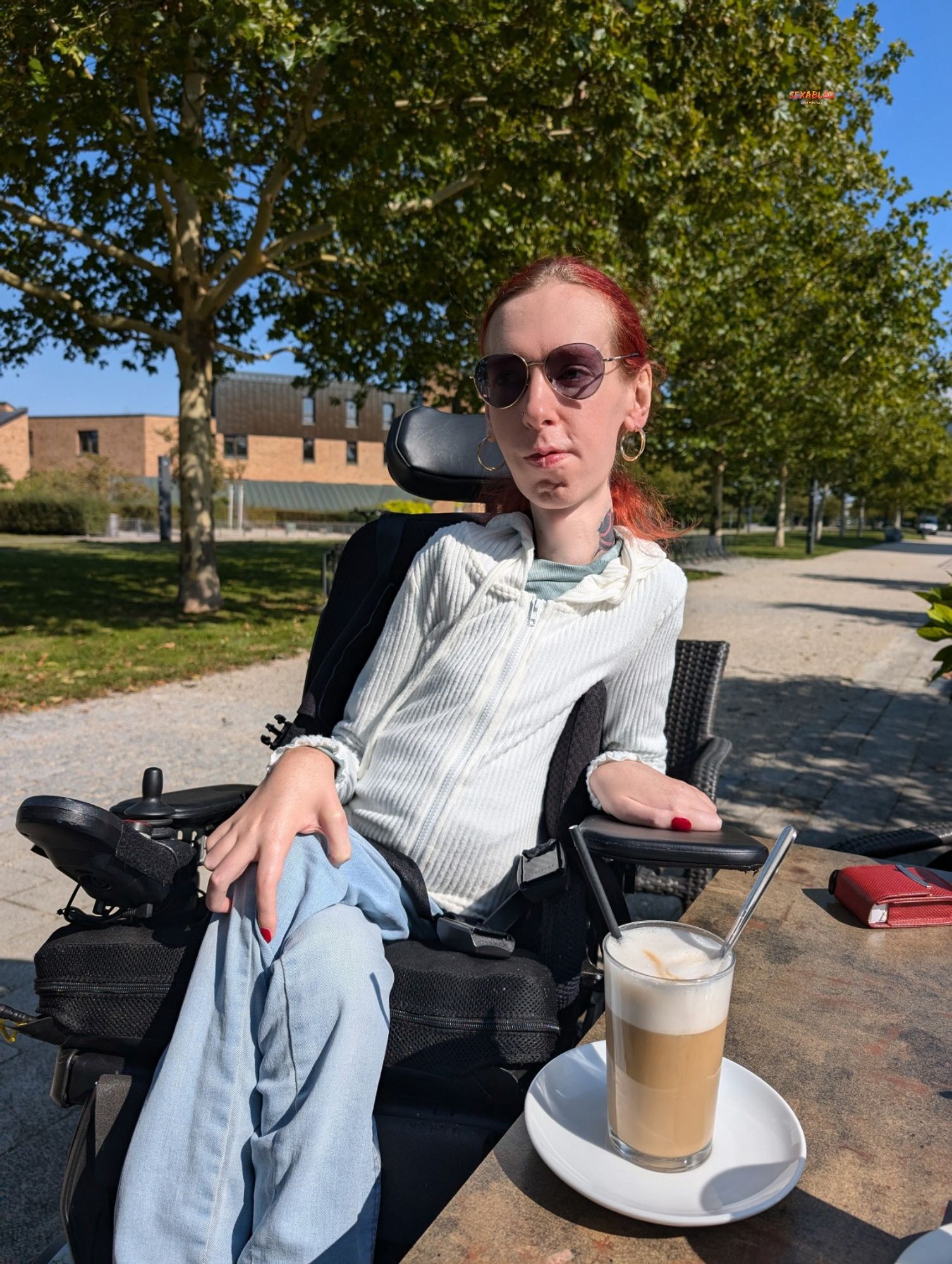 Ich sitze in einem Park, im Hintergrund sind Bäume und ein Weg zu sehen, vor mir auf dem Tisch steht der Kaffee. Ich trage ein weißes Jäckchen und eine hellblaue Jeans. Meine Lippen werden von einem leichten Lächeln umspielt und mein Blick geht in die Ferne.