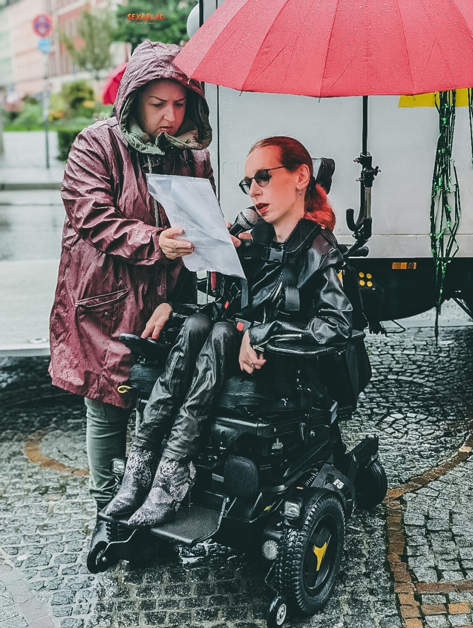 Slavica und ich stehen am Gärtnerplatz in München vor dem Wagen des Slutwalks. Es regnet stark. Sie trägt eine rote Regenjacke mit Kapuze und blickt konzentriert auf die Rede, die sie für mich in ihrer rechten Hand hält, während ich spreche. In ihrer linken Hand hält sie mein Mikrofon.

Ich trage eine schwarze Lederjacke, eine schwarze Lederhose und Stiefel mit einem Schlangenhautmuster. Meine roten Haare sind wetterbedingt zu einem Zopf gebunden. Ein roter Regenschirm, der mit einer Halterung an meinem Rollstuhl befestigt ist, schützt mit zumindest etwas vor dem Regen.