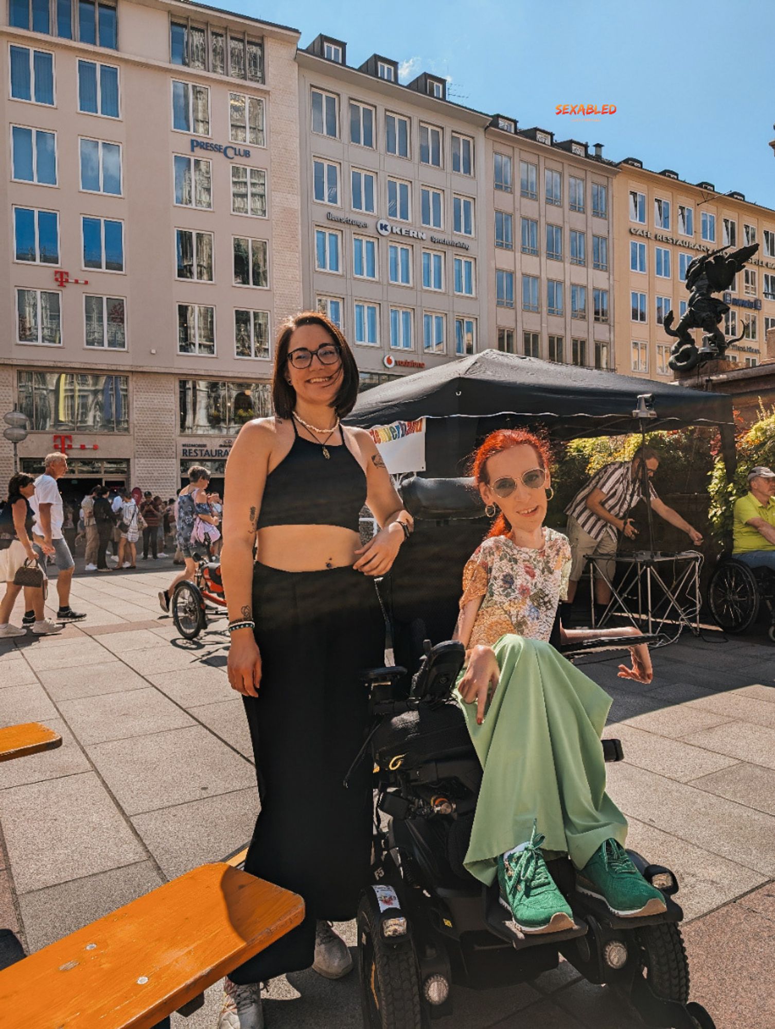 Franzi und ich stehen mitten auf dem Marienplatz. Wir lächeln in die Kamera. Hinter uns sieht man einige Menschen, Geschäfte und der Pavillon für die Technik der Demo.

Franzi trägt ein schwarzes bauchfreies Top und eine schwarze Stoffhose mit weißen Sneakern. Ich trage eine halb durchsichtige Bluse mit buntem Blumenmuster, eine hellgrüne Stoffhose und dunkelgrüne Sneaker.