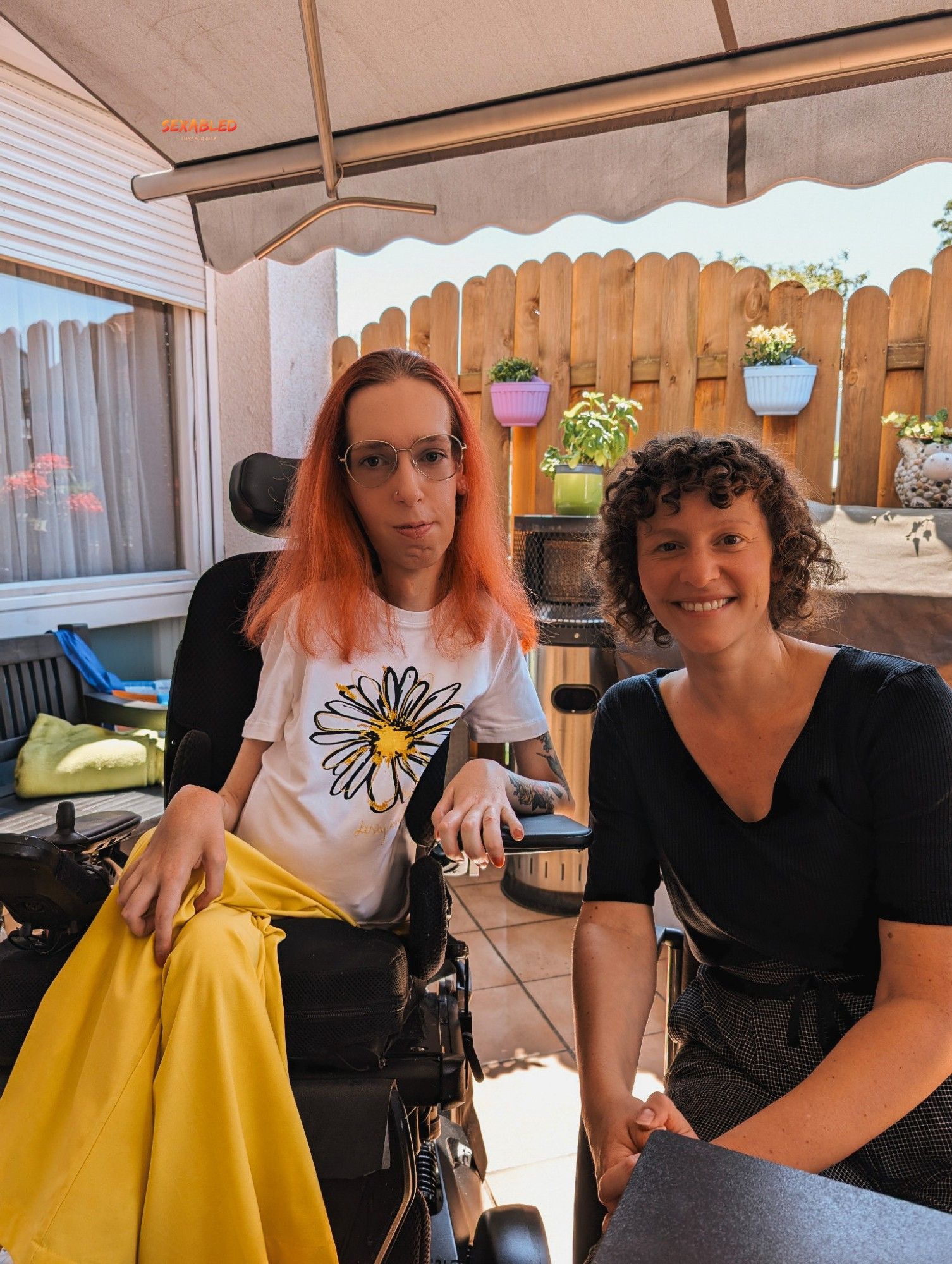 Ich sitze zusammen mit Ekaterina auf meiner Terrasse. Wir sitzen nebeneinander und lächeln in die Kamera. Ich trage ein weißes Shirt mit einer Blüte auf der Brust und gelbe Stoffhose. Meine leicht rötlichen Haare sind geöffnet und fallen über meine Schultern. Ekaterina trägt eine schwarze Bluse, eine schwarz weiß gemusterte Stoffhose und mittellanges lockiges braunes Haar.