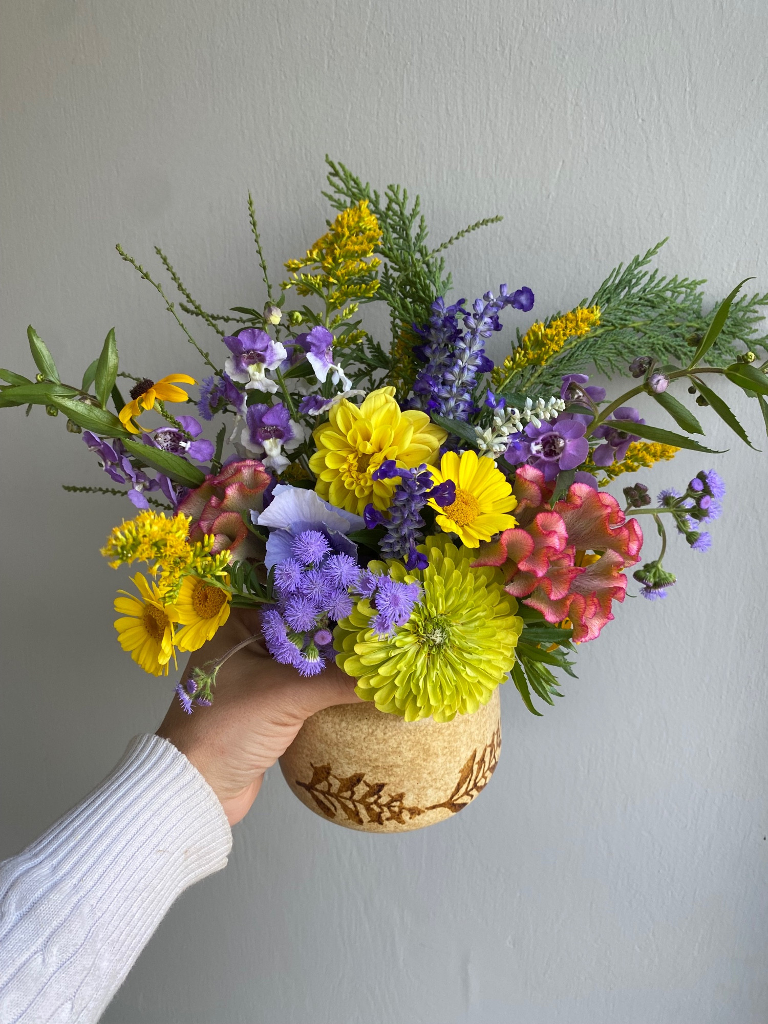 A hand holding a tan ceramic vase with brown leaf details which contains a colorful bouquet of ageratum, pansies, zinnias, celosia, painted daisies, angelonia, black eyed Susans, white vervain, salvia, and cypress clippings in front of a plain gray wall.