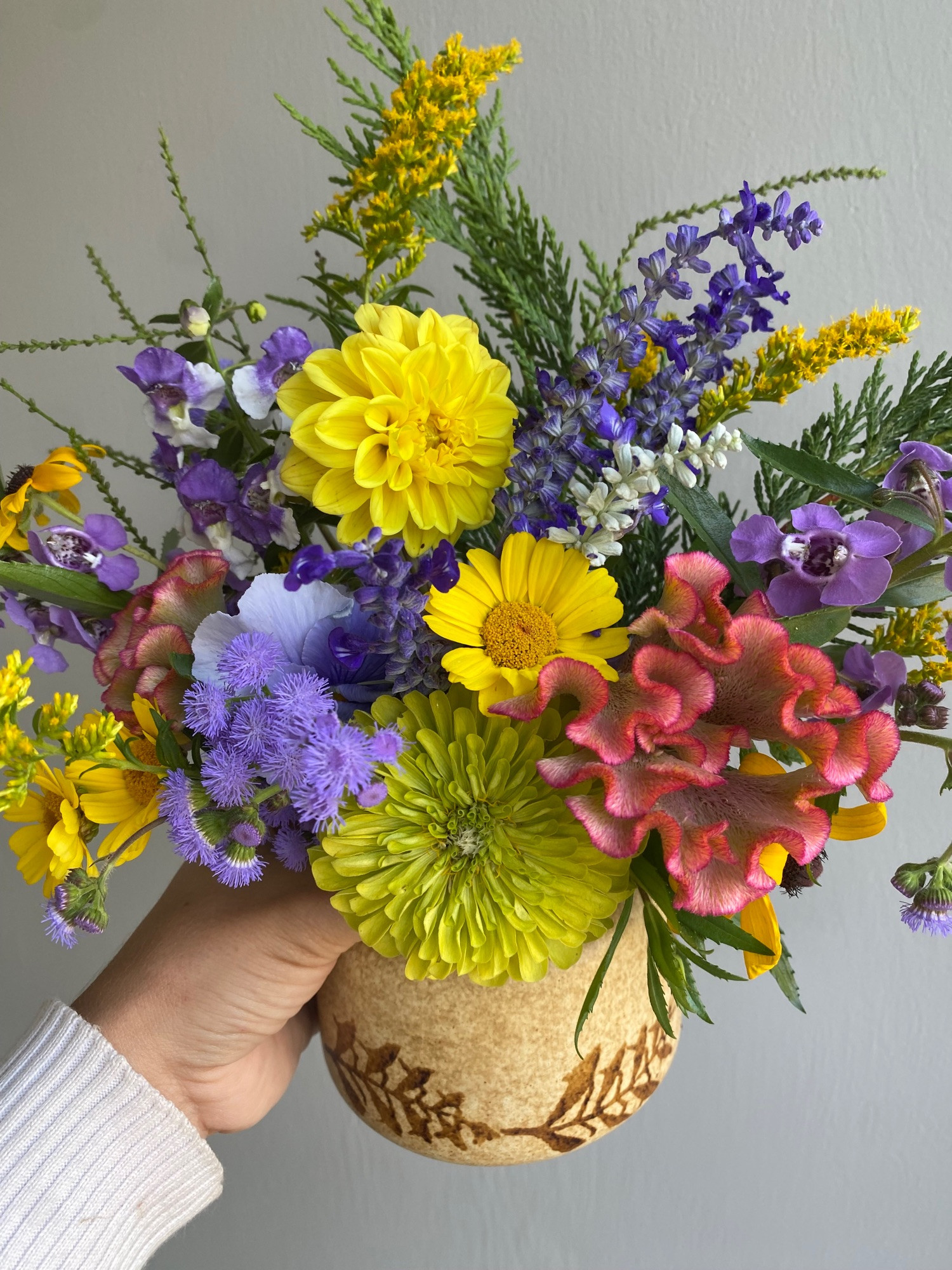 Closer look at the same hand holding a tan ceramic vase with brown leaf details which contains a colorful bouquet of ageratum, pansies, zinnias, celosia, painted daisies, angelonia, black eyed Susans, white vervain, salvia, and cypress clippings in front of a plain gray wall.