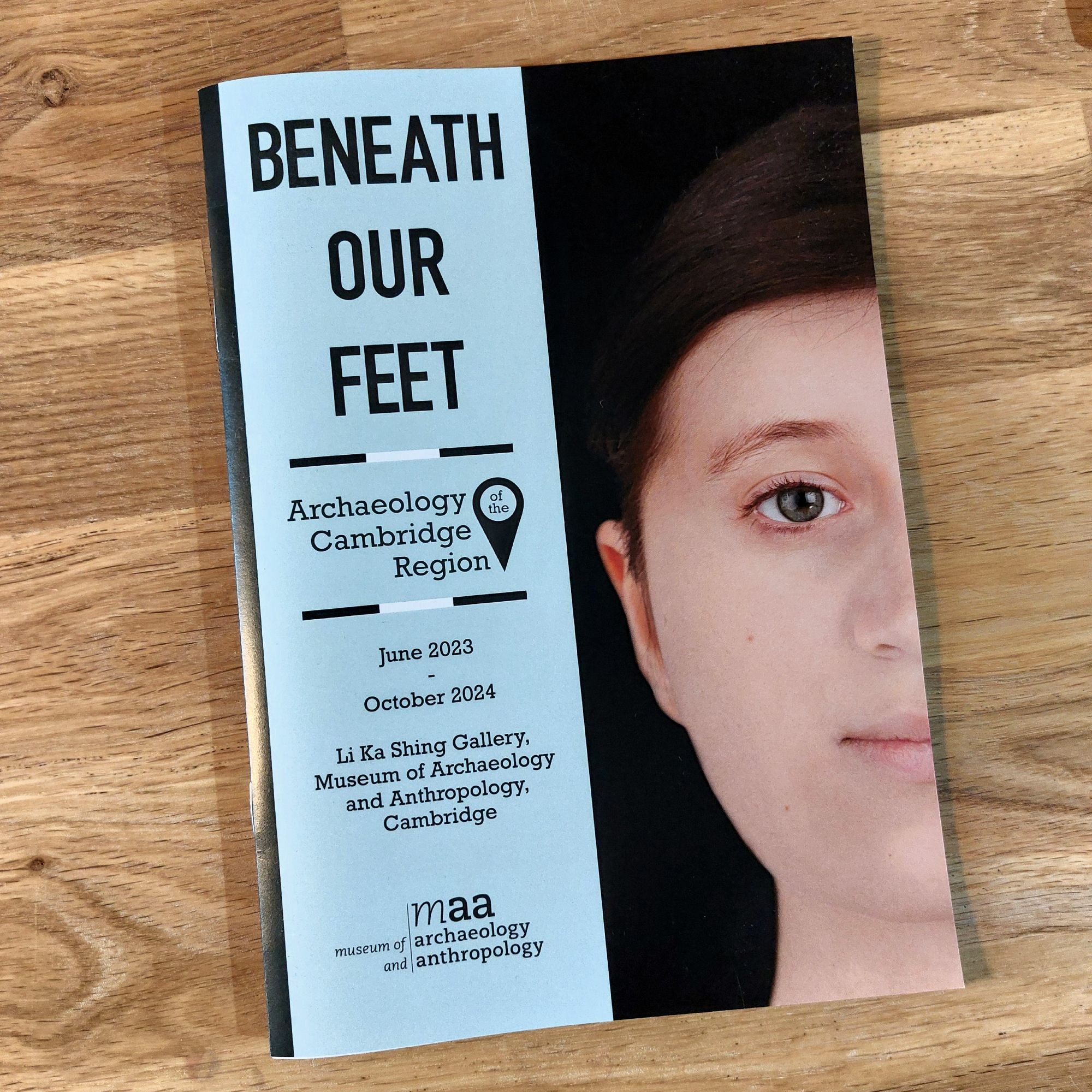 Photograph of the exhibition booklet for the 'Beneath our Feet: Archaeology of the Cambridge Region' exhibition at the MAA in Cambridge. The cover shows a facial reconstruction of a early medieval female found buried at Trumpington.