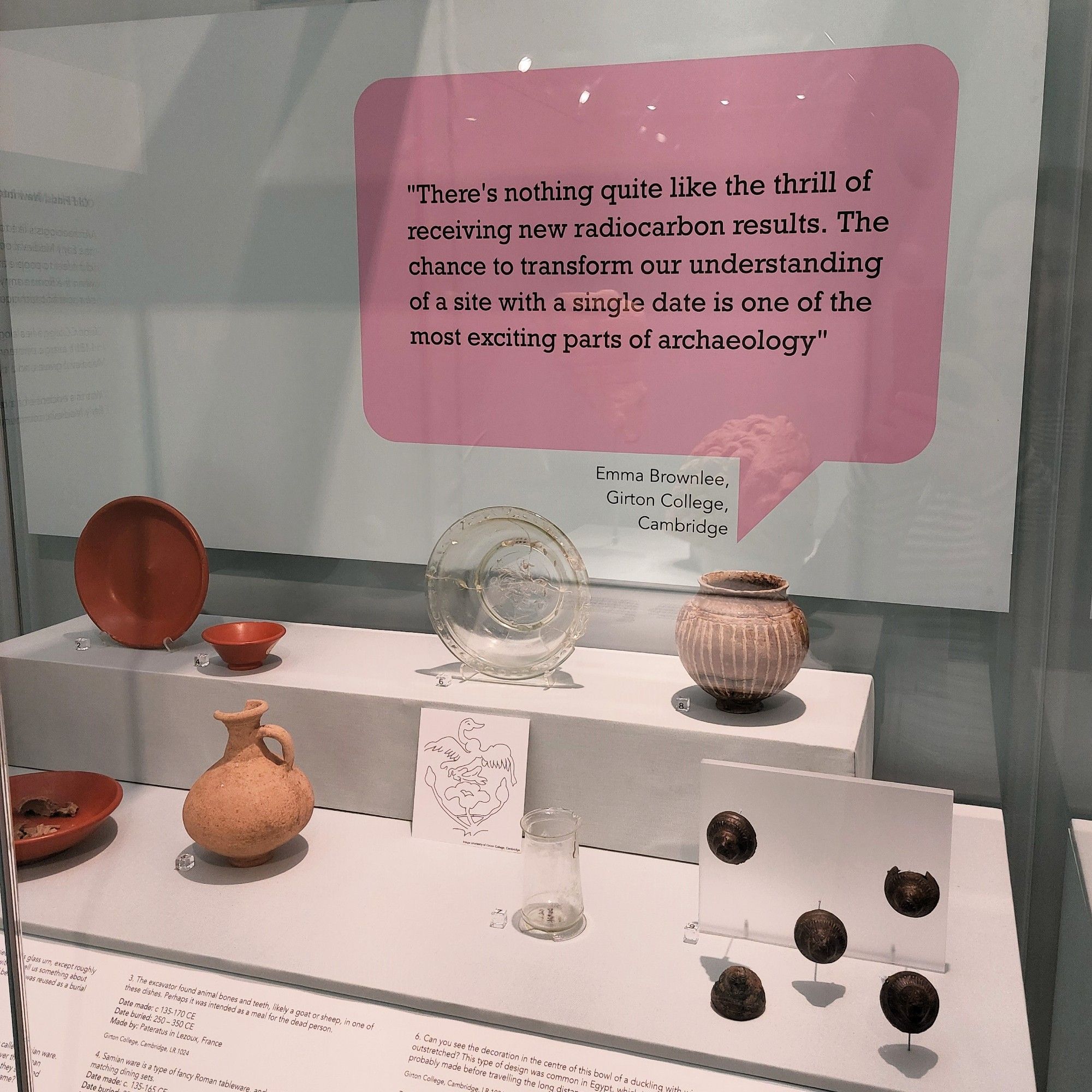 A photograph of a display of Roman and early medieval artefacts found at Girton College. The display includes a quote from Emma Brownlee.