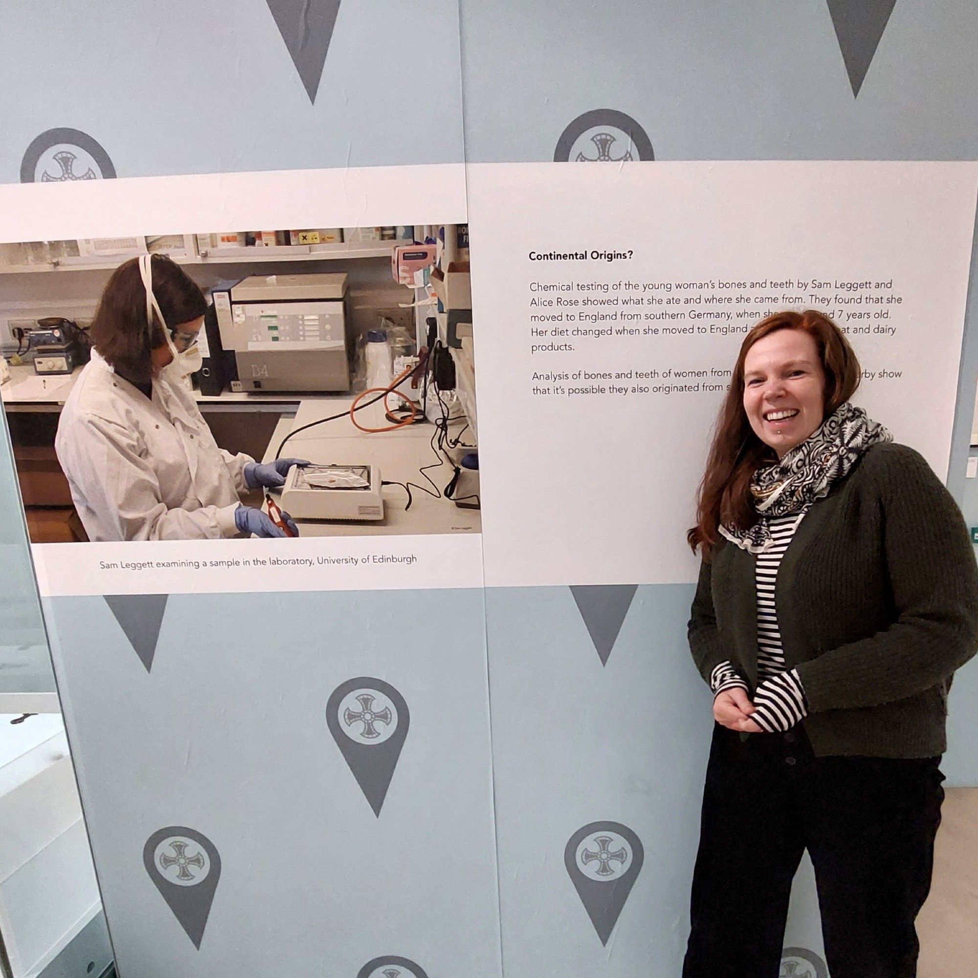 Photograph of the author, Alice Rose, standing in front of an information board in the 'Beneath our Feet' exhibition. The board describes scientific work that Alice Rose and Sam Leggett carried out on the Trumpington burial.