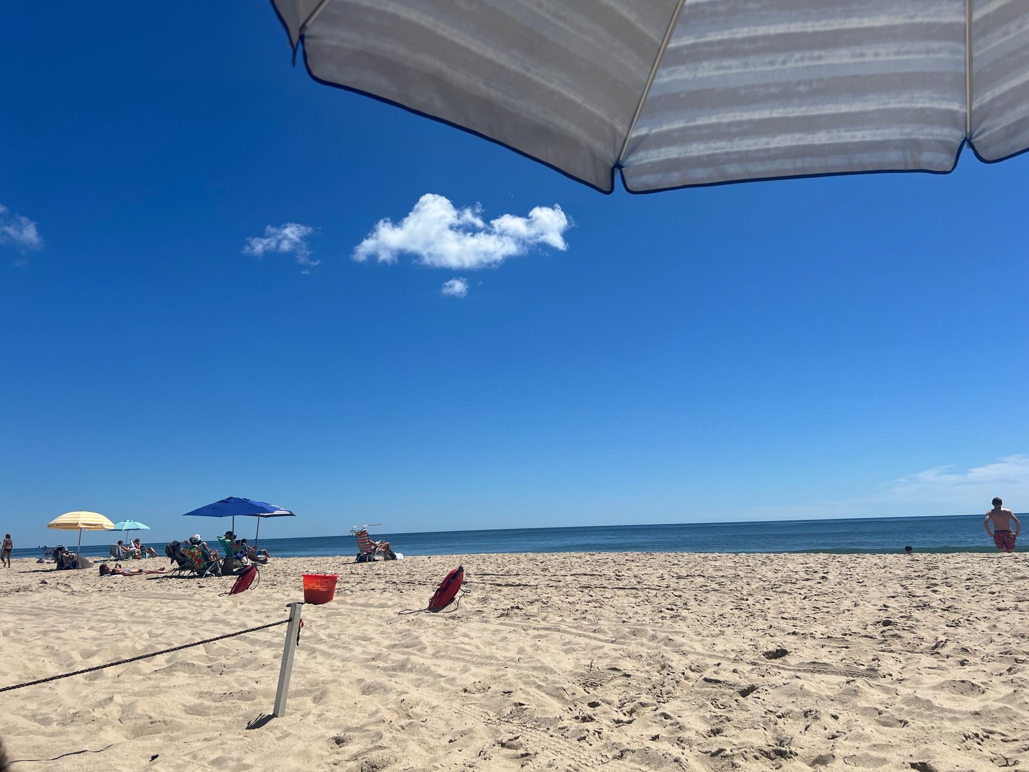 The beach. It's beautiful. One impossibly playful cloud in a richly blue sky. A blue beach umbrella peeks into the frame on the upper right; more umbrellas and a bather are in the middle distance. A wooden spike and rope mark the province of the lifeguards.