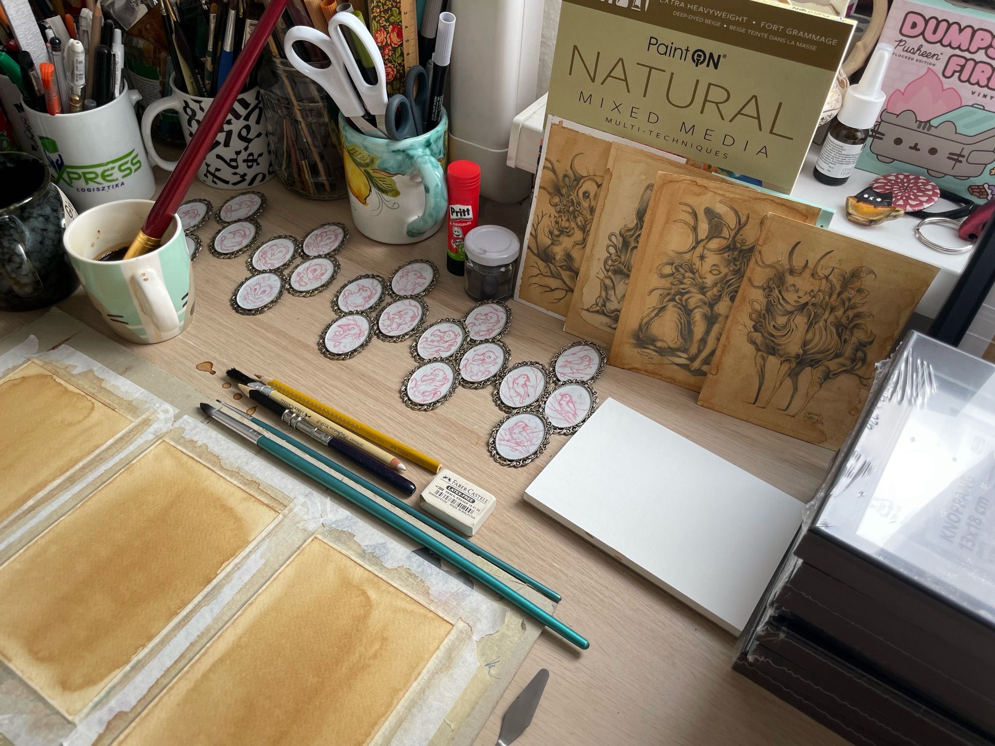 Workspace on a table. Coffee stained papers, graphite drawings, sketched up brooches, art supplies.