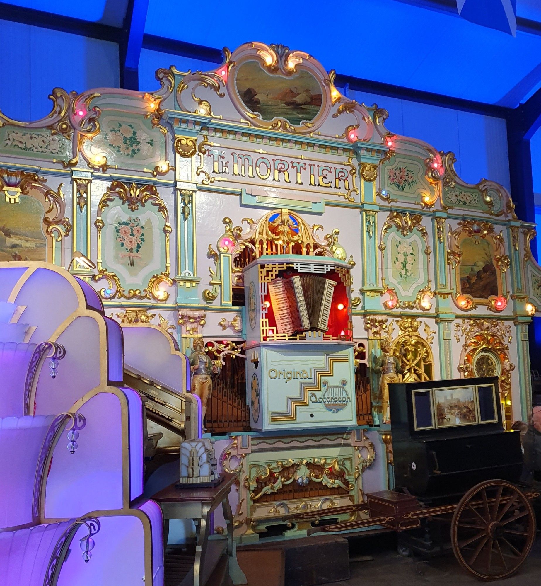 A huge duck egg blue Th Mortier fairground organ with much baroque gold ornamentation and an accordion mounted on the front, with art deco lettering below labelling it as "original accordion". At the front left is a Wurlitzer organ seen from the side, and to the right of the Mortier is a wagon-amounted barrel organ