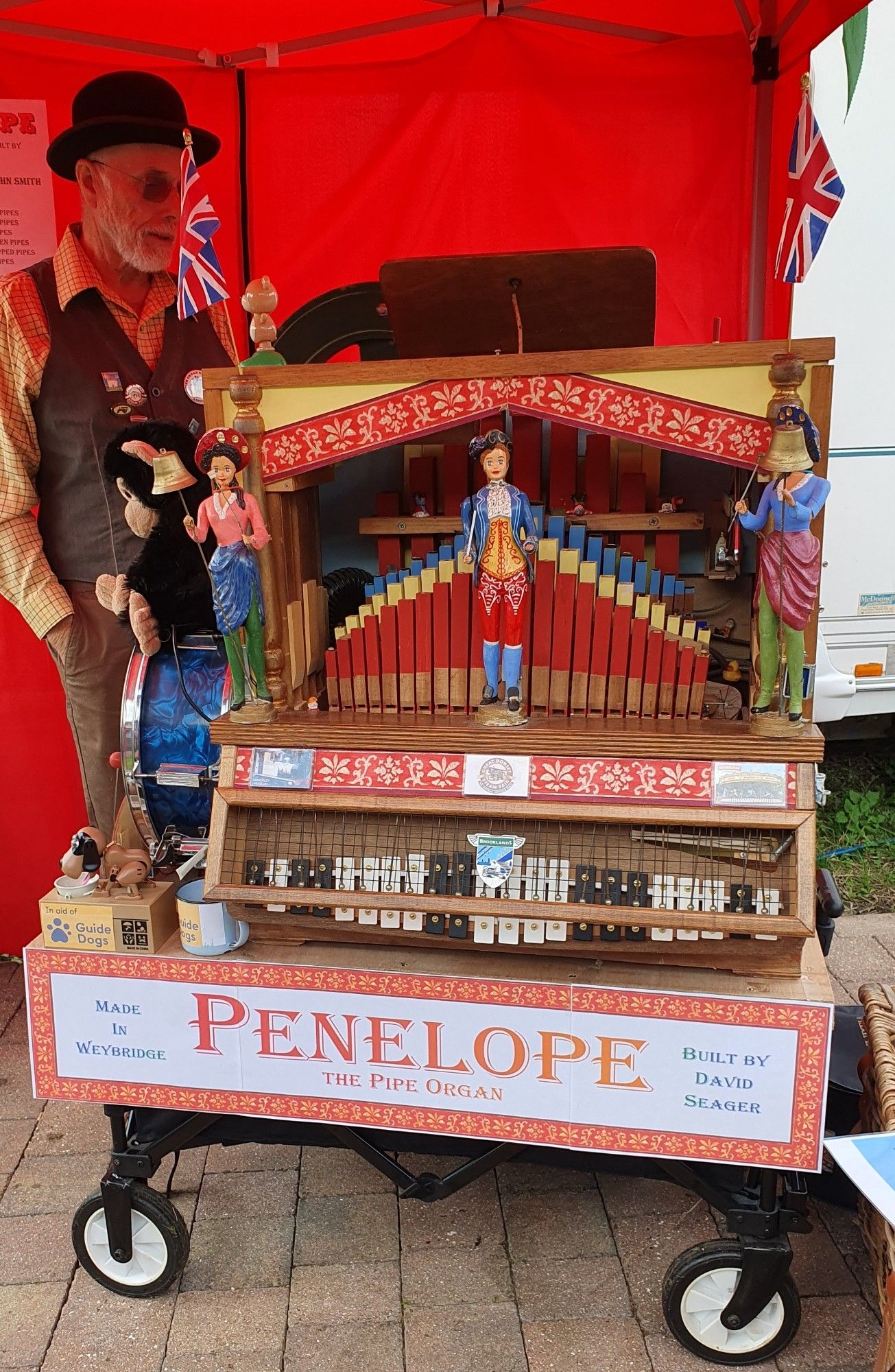 Penelope the pipe organ, with a label proclaiming that she was made in Weybridge by David Seager. She is a fairly small pipe organ with full glockenspiel across the bottom and three doll figurines standing in front of the organ pipe display. Beside her stands an elderly white man wearing a bowler hat, dark glasses, and a brown waistcoat. A collection box for a Guide Dog charity sits beside the organ.