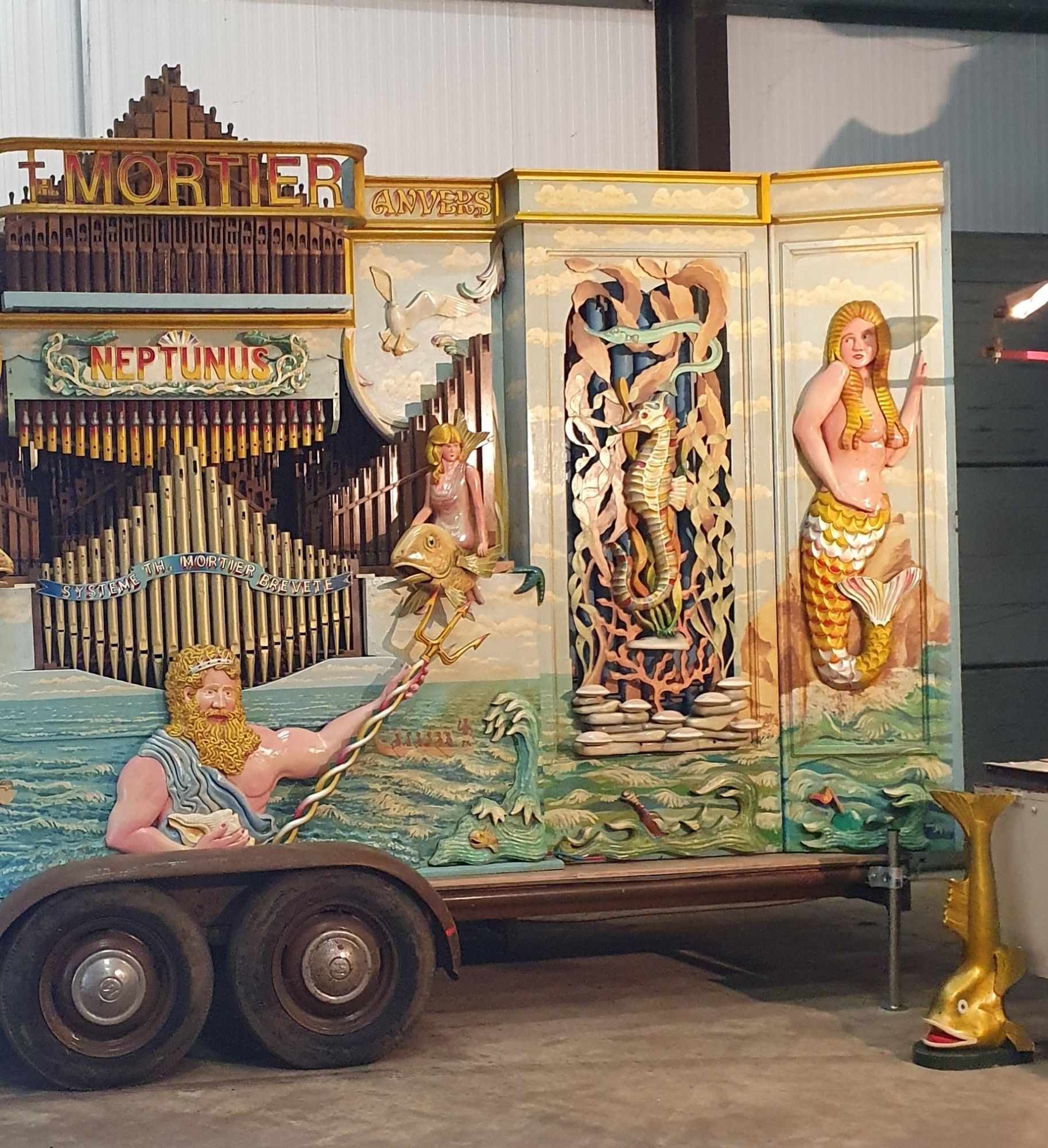 A very elaborate Mortier fairground organ labelled as "Neptunus". A figure of Neptune holding his Triton is on the front centre bottom panel, bi's head at the bottom centre of a row of organ pipes, and to the right side a smaller figurine is mounted on a fish. To the right of that is an elaborately carved seahorse surrounded by kelp, and to the right of the seahorse panel is a relief of a mermaid with a golden tail and long blonde hair covering her bare breasts. In front of the organ is a relatively smaller lectern shaped like an open-mouthed golden fish, with its tail as the top. The organ is mounted on a long trailer