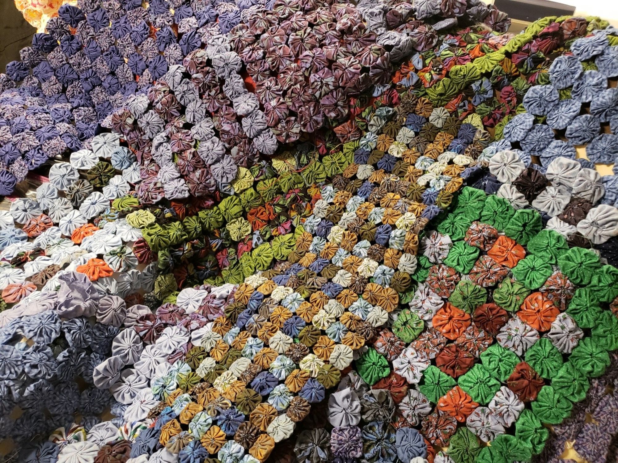 Thirteen or fourteen table runners of multicolored cotton cloth, all found at Goodwill for $1.20 to $1.90 each, now overdye with royal blue. Each runner is made of a rectangular grid of circular puffy fabric yoyos, each one joined on the sides by hand-stitching.

In this photo, they look like a hilly forested landscape seen from the air.