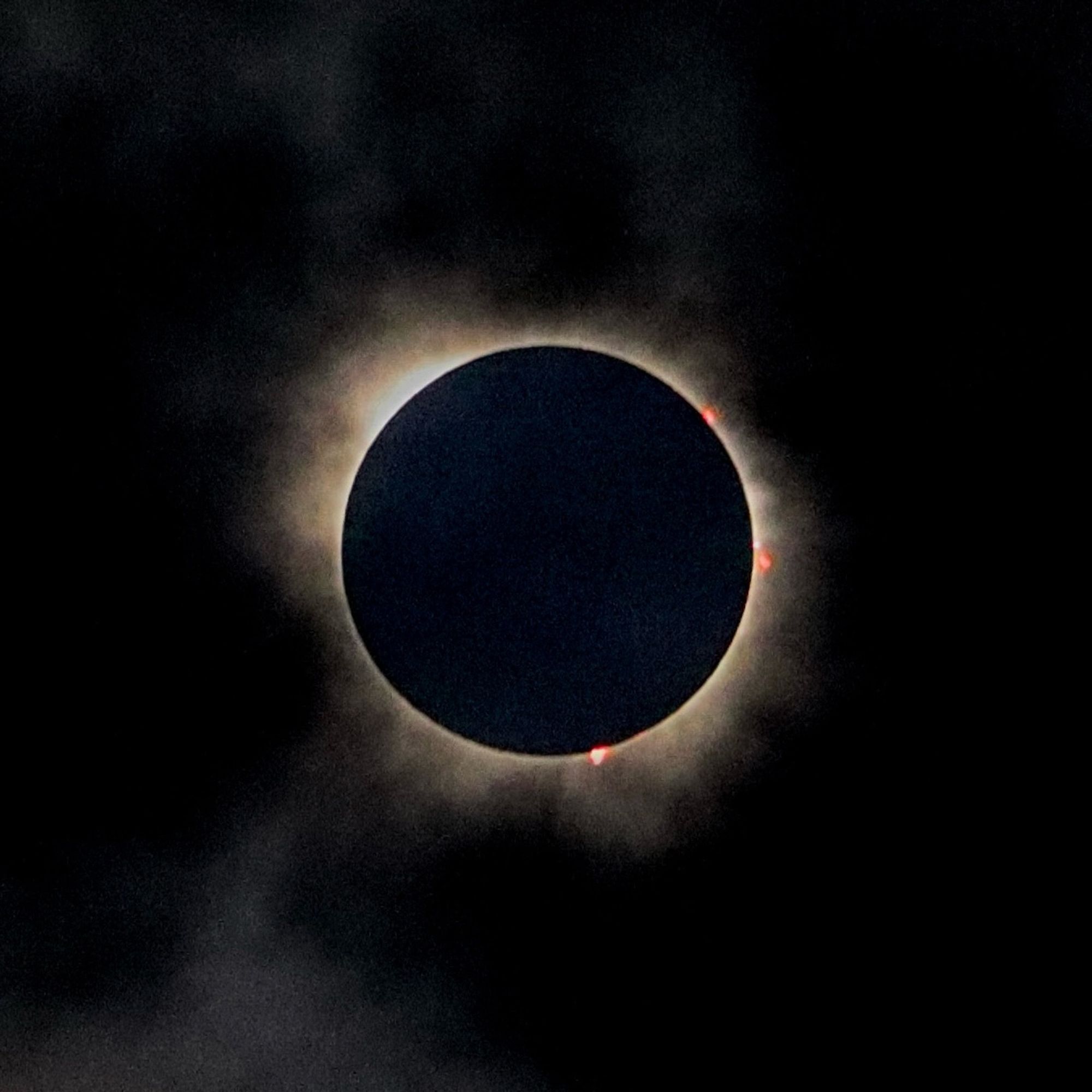 A cloudy view of the solar eclipse during totality, featuring flame-like pink prominences leaping out from the edge of the disc