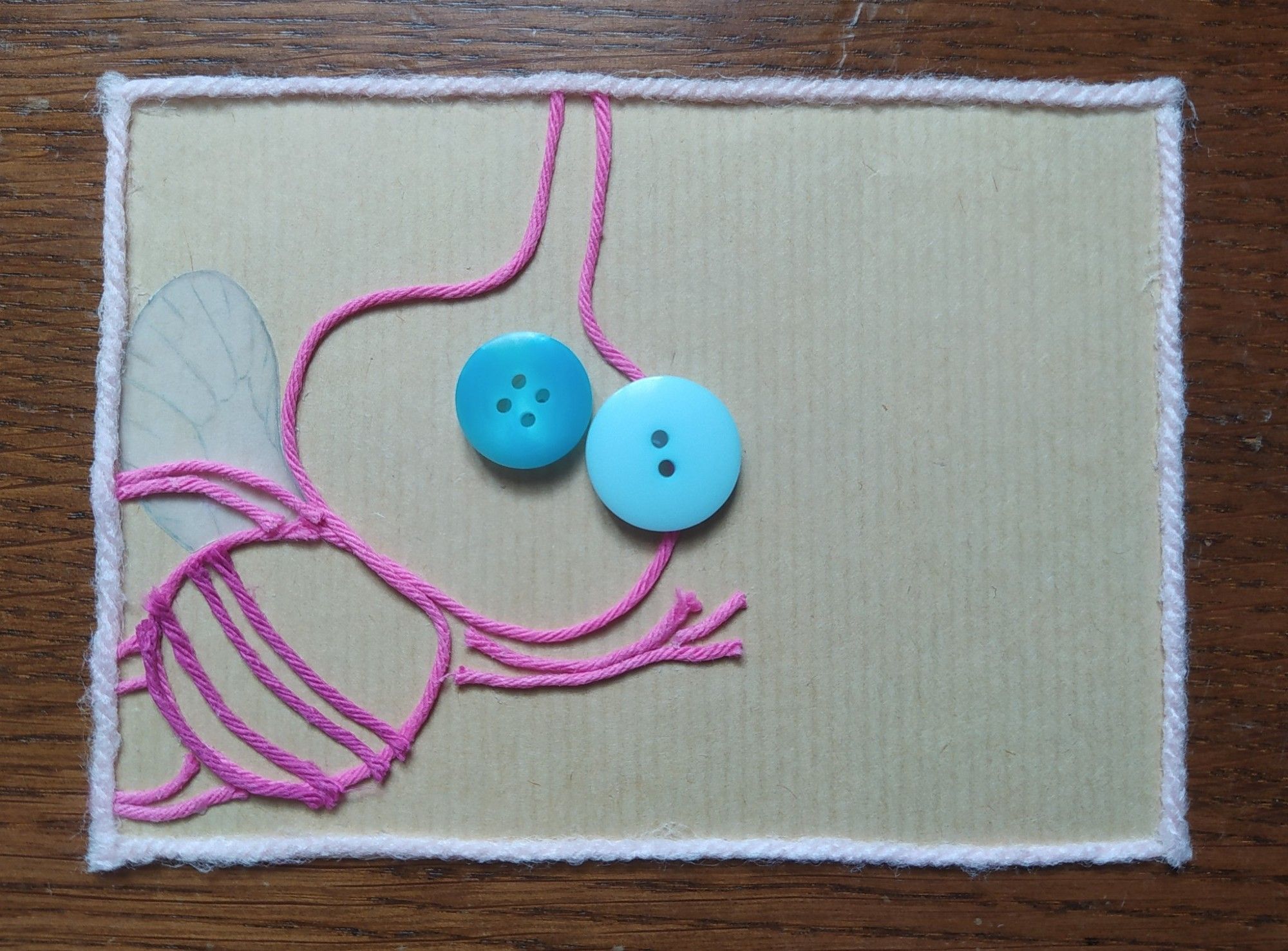 Art of a Winged Pikmin waving it's hand and looking toward you.
The outline of the Pikmin is made of pink wool thread. It's eyes are represented with two blue buttons of different shades and size (it's left eye is a big pale blue button with two holes while it's right eye is a more vibrant blue button with four holes and is slightly smaller). It's wing is made of tracing paper that's been drawn onto with pencils.

The background is a piece of brown kraft paper and there is a thicker pale pink wool thread on the edges to create a frame.