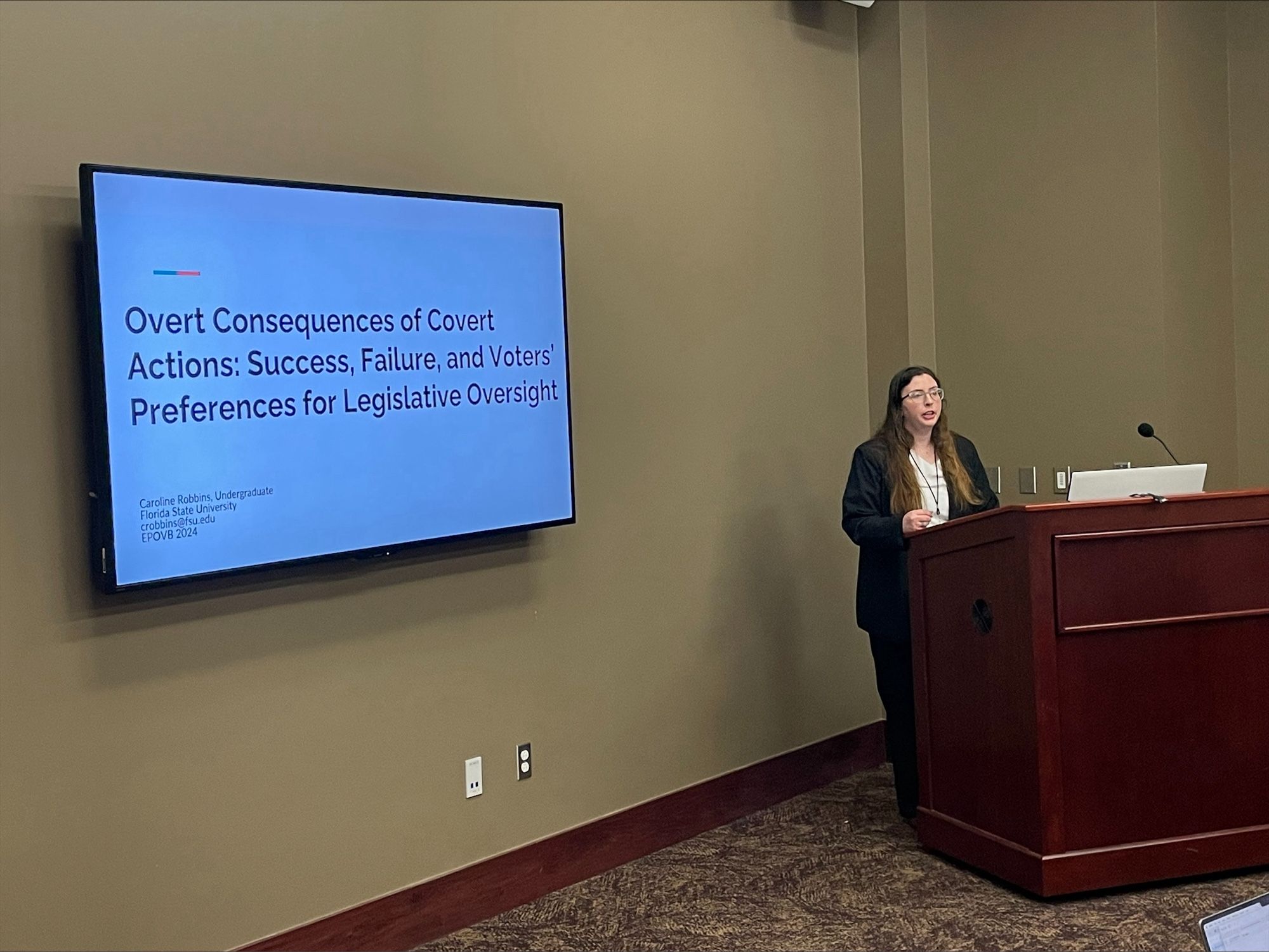 Caroline standing to the right at a dark wood podium presenting with a television screen on the left displaying the title of the article “Overt Consequences of Covert Actions: Success, Failure, and Voters’ Preferences for Legislative Oversight.”