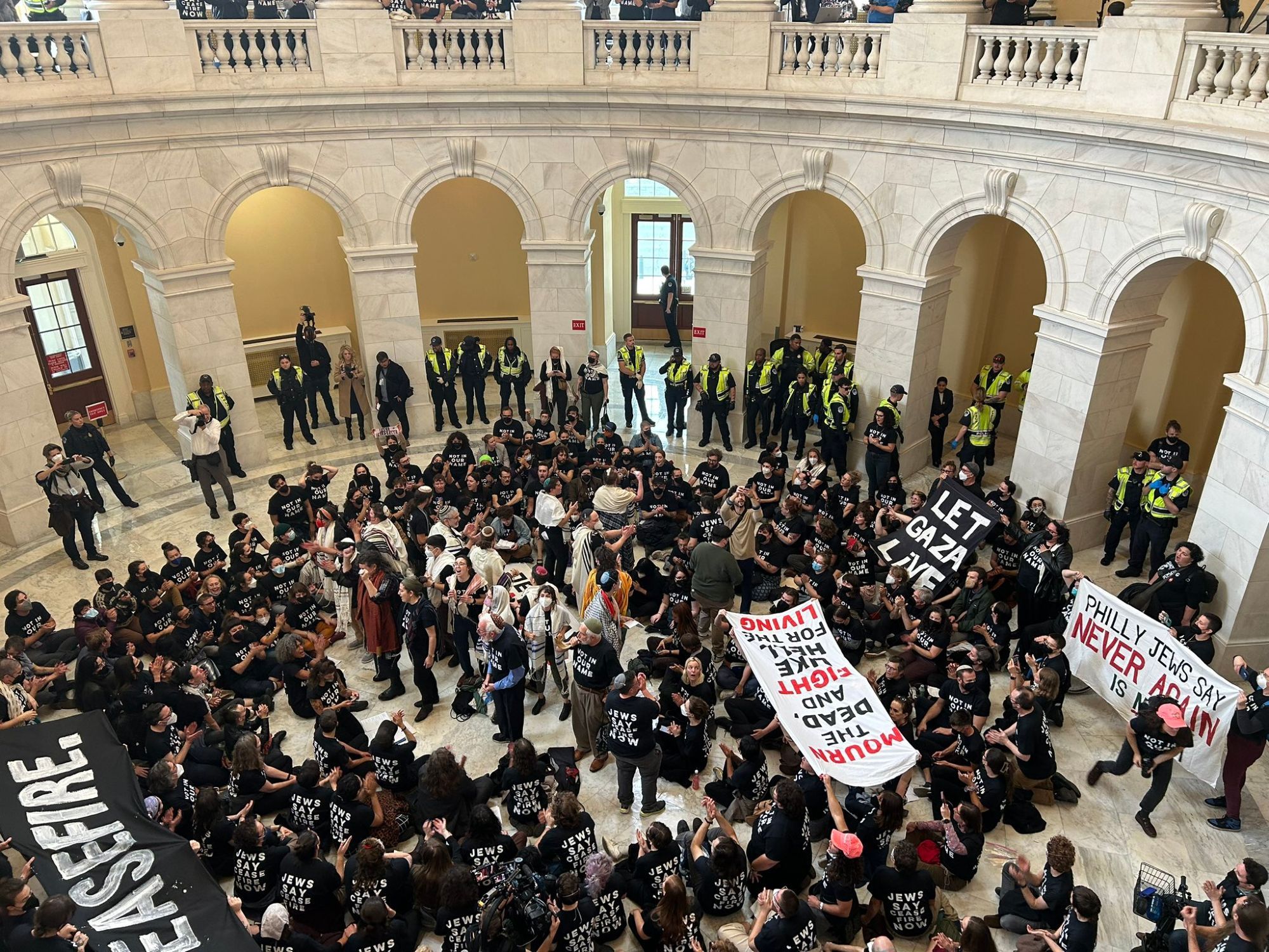 A picture of the Cannon rotunda. There is a large group of Jewish Voice for Peace protesters in black shirts holding signs that say things like "Let Gaza live" "ceasefire" "Philly Jews say Never Again Is Now" "Mourn the dead and fight like hell for the living." They fill the middle of the rotunda, many are sitting down. There are folks on the edges in yellow vests who I believe are law enforcement but may be marshals/observers/press? My eyes aren't that great.