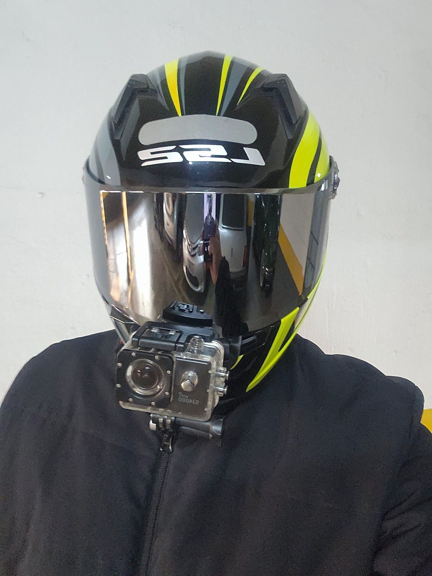 Uma foto minha utilizando um capacete preto com detalhes verde limão e cinza com uma viseira espelhada e uma câmera estilo Gopro acoplada