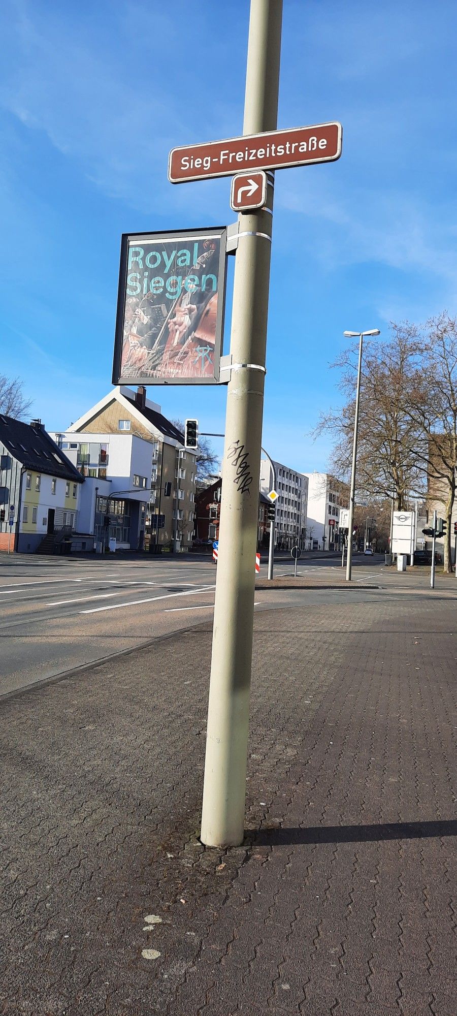 Blick auf eine leere Straße mit Häusern am Rand. Auf dem Gehweg ein langer Pfeiler, an dem ein Poster mit dem Titel "Royal Siegen" befestigt ist sowie ein brauner, weiß beschriebener Wegweiser mit der Aufschrift "Sieg-Freizeitstraße".